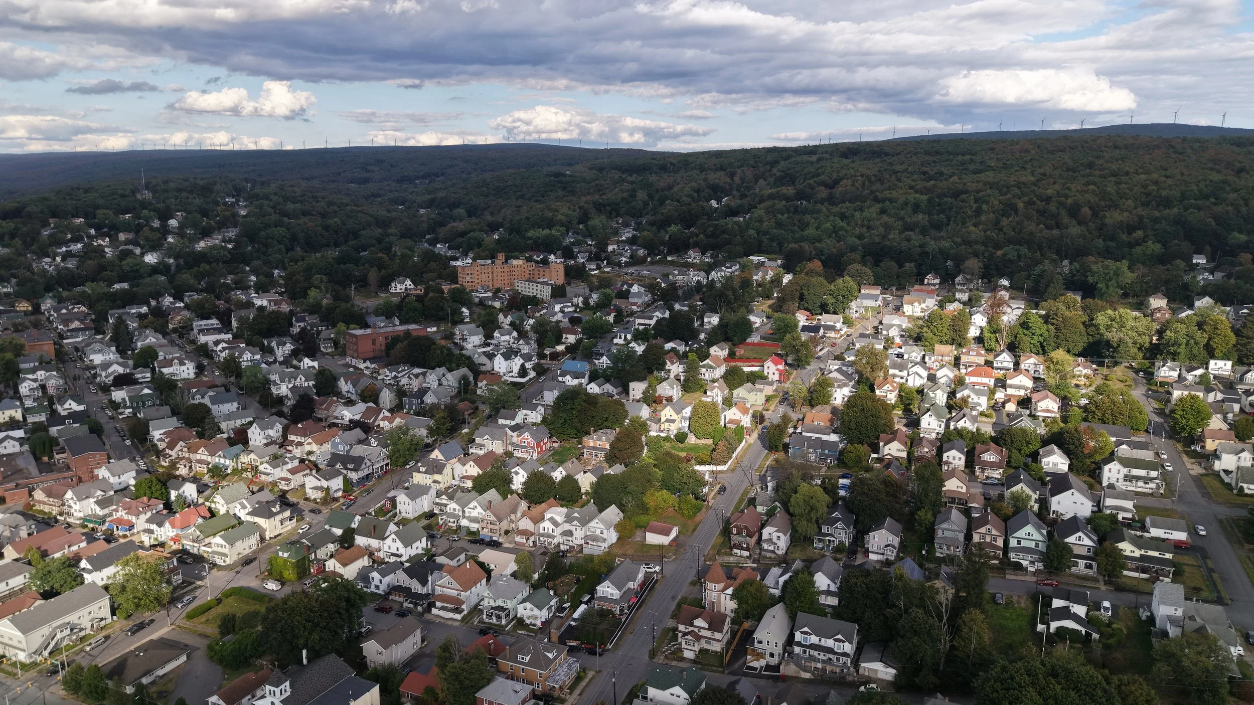 Aerial view of a residential neighborhood with houses, streets, and green trees, bordered by a forested hillside and wind turbines on a distant ridge under a cloudy sky.