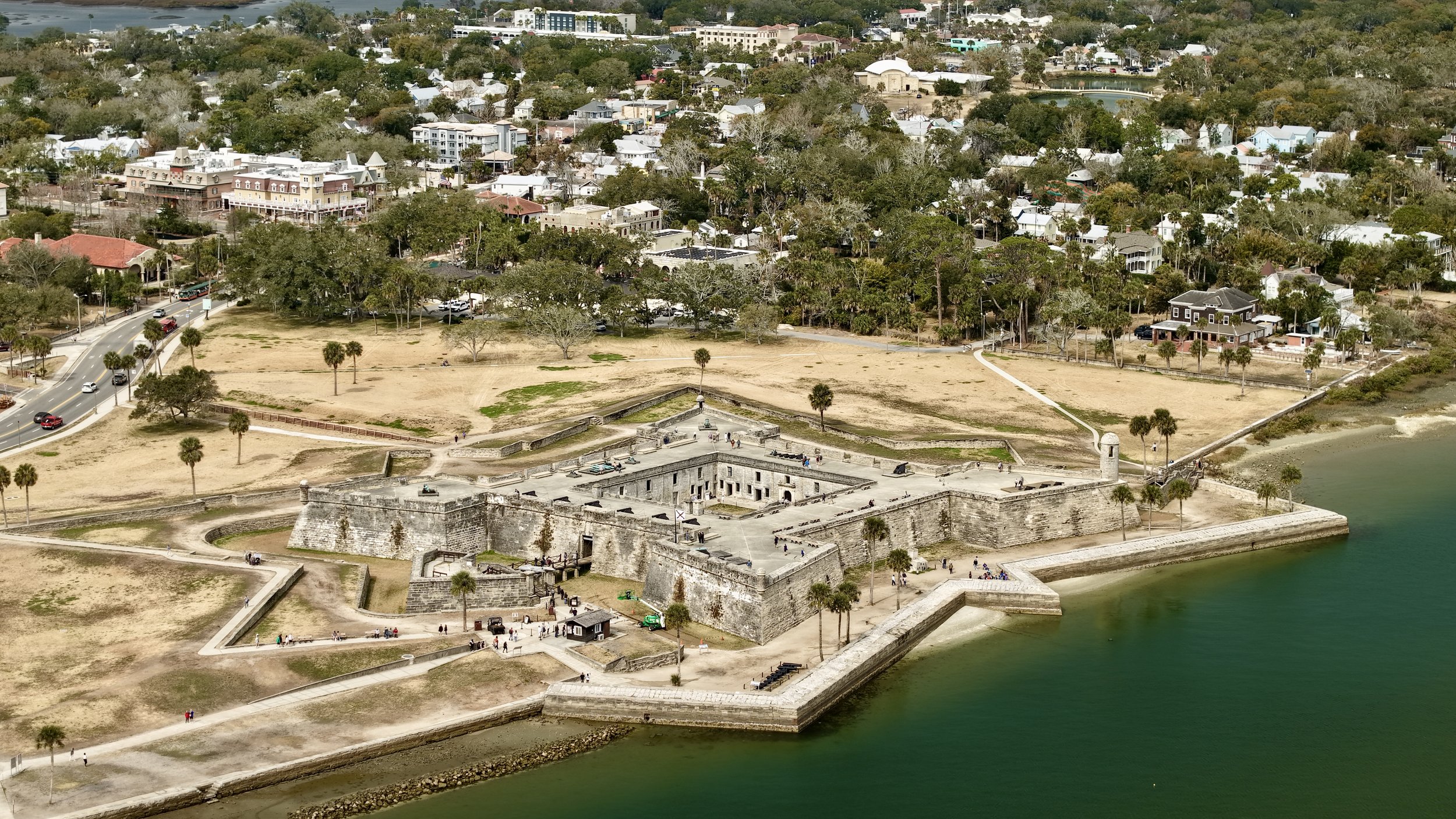 Aerial view of a historic fort located by the water, with a large open grassy area and city buildings in the background.