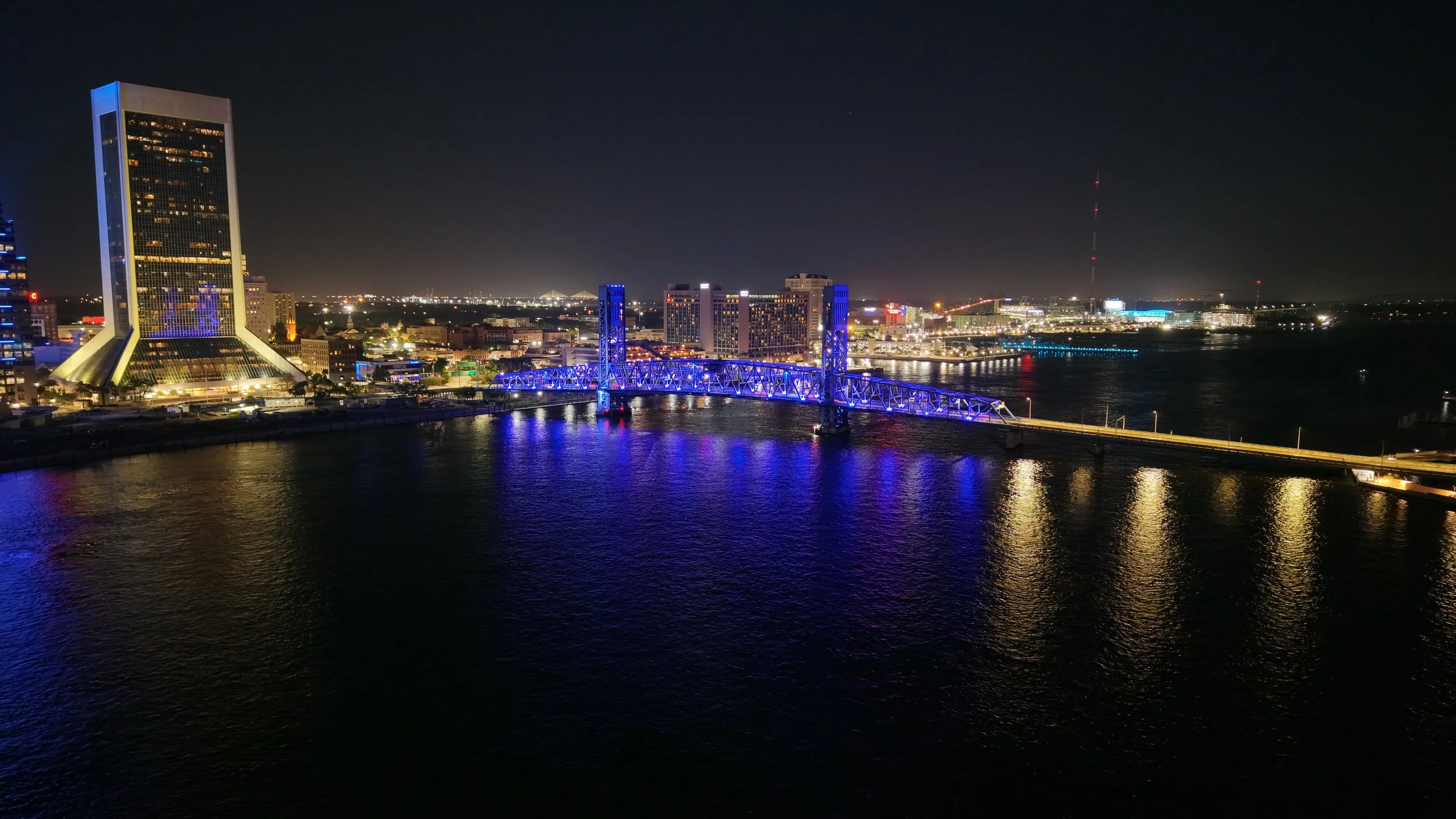 Night view of a city skyline with a lit-up bridge reflecting on the water, including tall buildings and a radio tower in the distance.