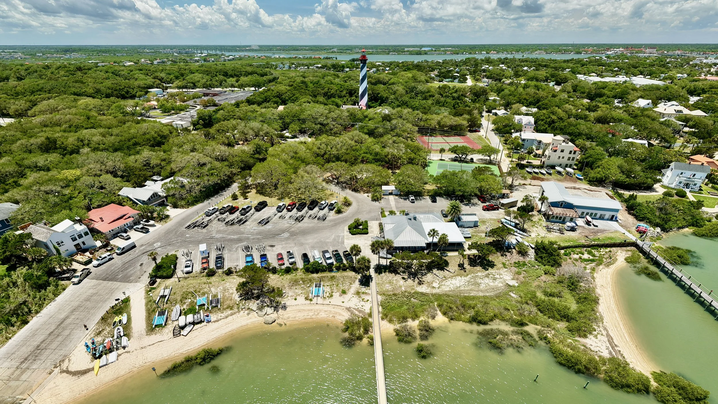 Aerial view of a coastal area with a lighthouse, tennis courts, parking lots, boats, and a beach with boats and kayaks, surrounded by greenery and water.