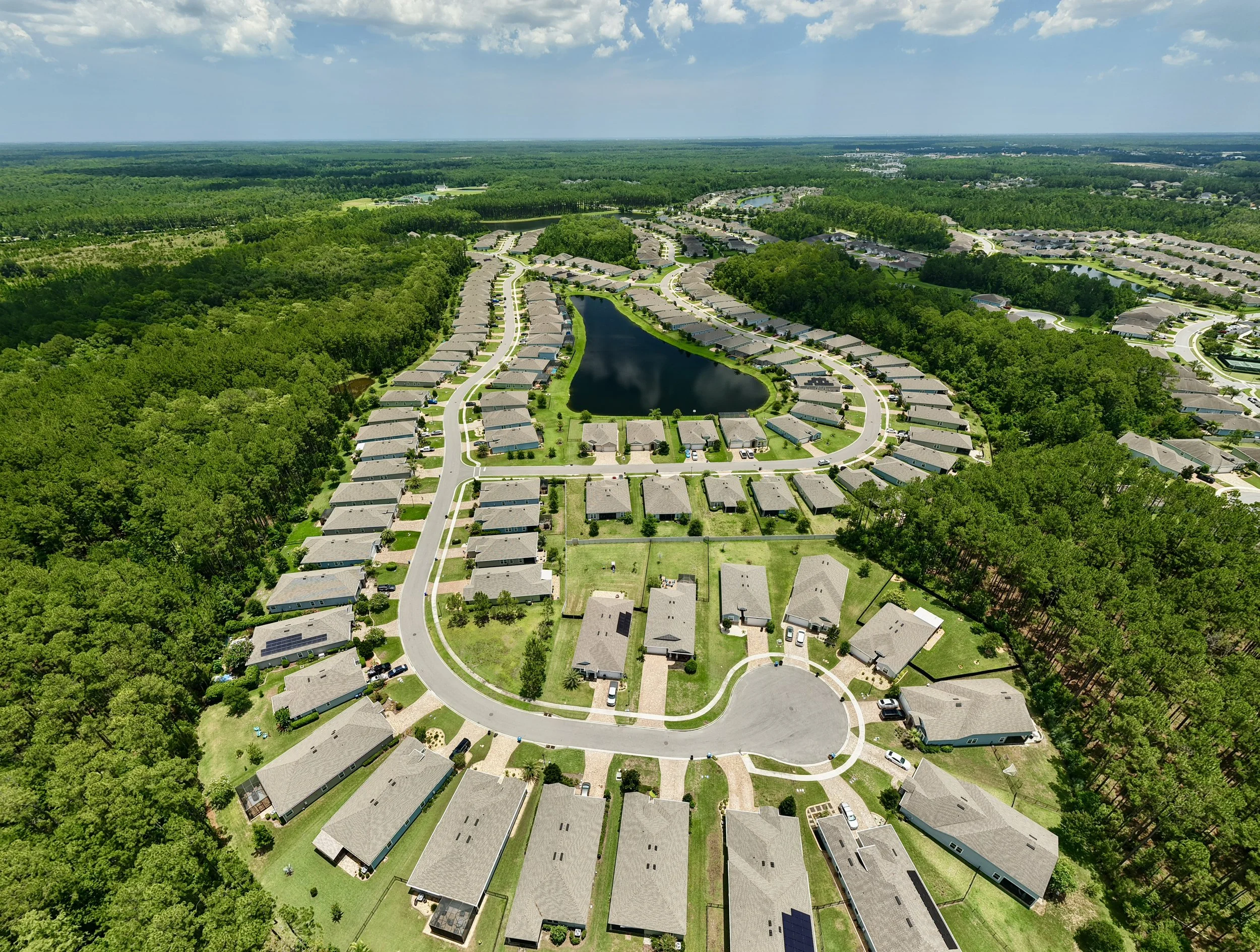 Aerial view of a suburban neighborhood with single-story houses, a small pond, surrounded by lush green trees and woodland, under a partly cloudy sky.