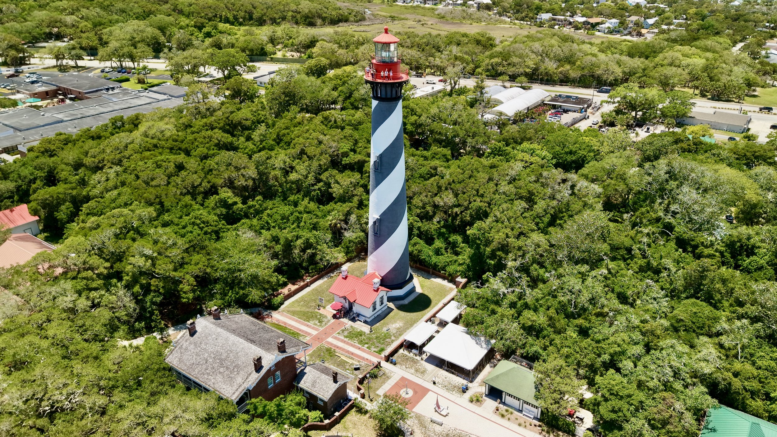 A tall lighthouse with a black and white spiral pattern, topped with a red roof, stands among dense green trees. There are small buildings nearby, and a parking lot with cars in the background. The scene appears to be in a coastal or tourist area.