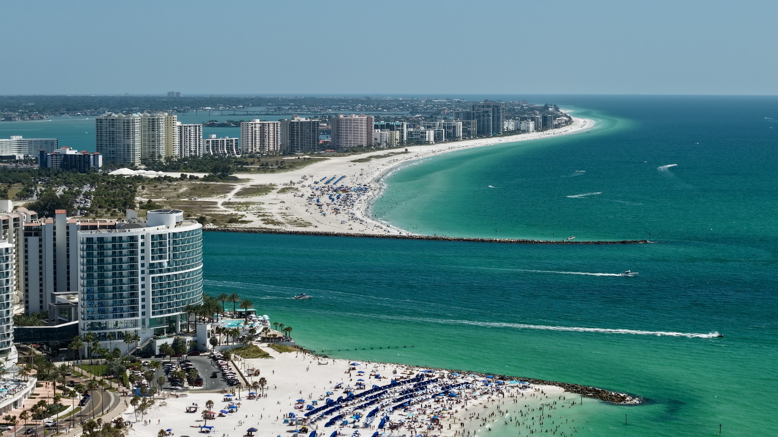 Aerial view of a coastal city with high-rise buildings, a sandy beach filled with umbrellas and people, and boats in the turquoise water.