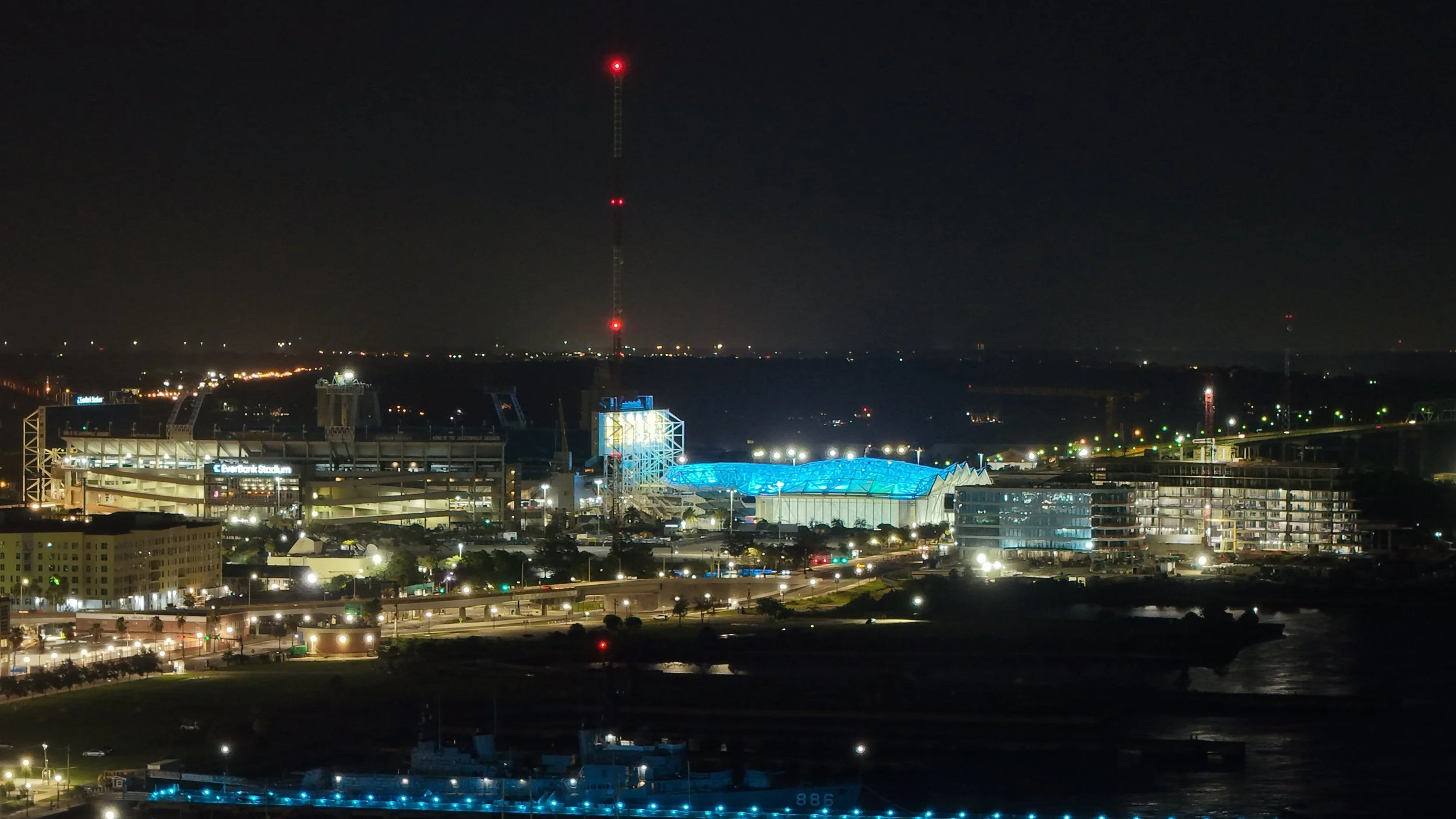 Nighttime cityscape featuring a stadium with illuminated blue roof, a tall radio tower with red lights, and various buildings with lights in the distance.