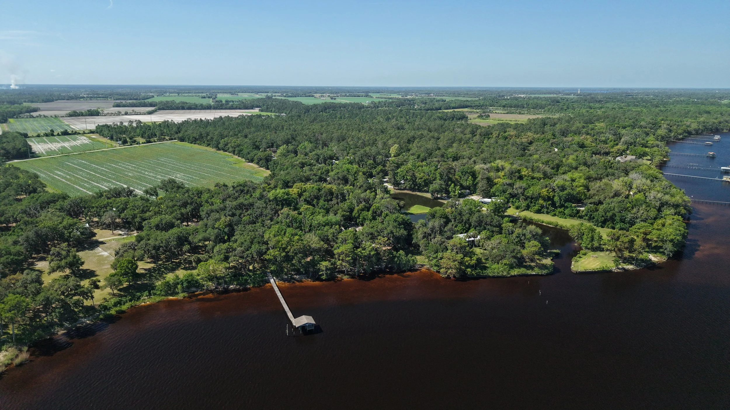 Aerial view of a rural landscape with farmland, trees, a river, and a marina with boats on the right side of the image.