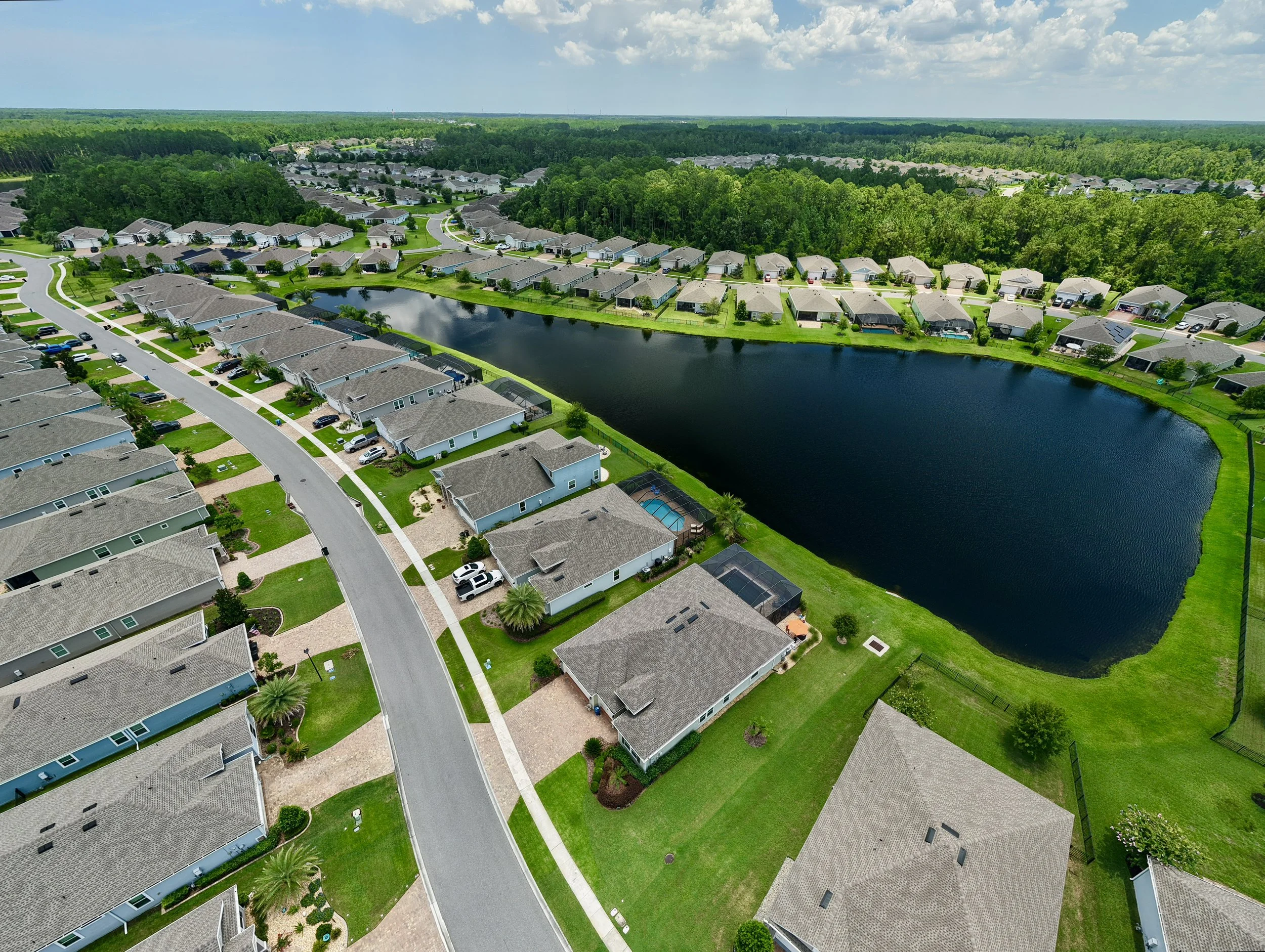 Aerial view of a suburban neighborhood with houses, a pond, green lawns, and surrounded by trees under a partly cloudy sky.