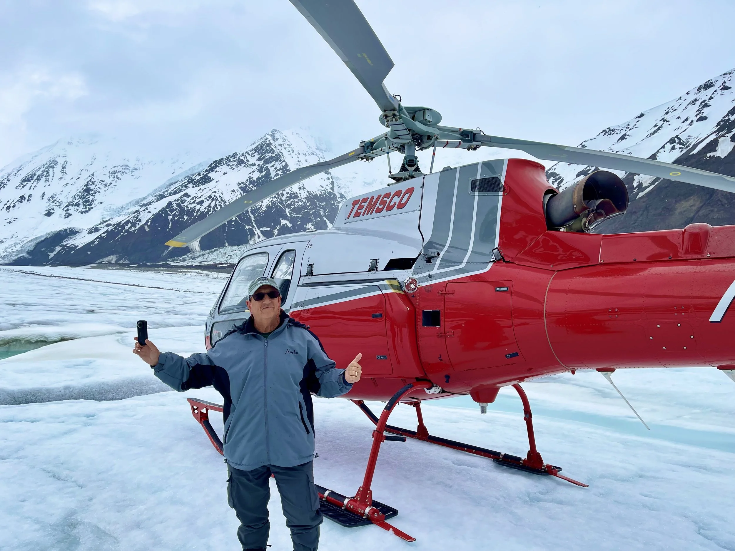 A man standing in snowy terrain in front of a red helicopter with mountains in the background, giving a thumbs-up and holding a phone in one hand.