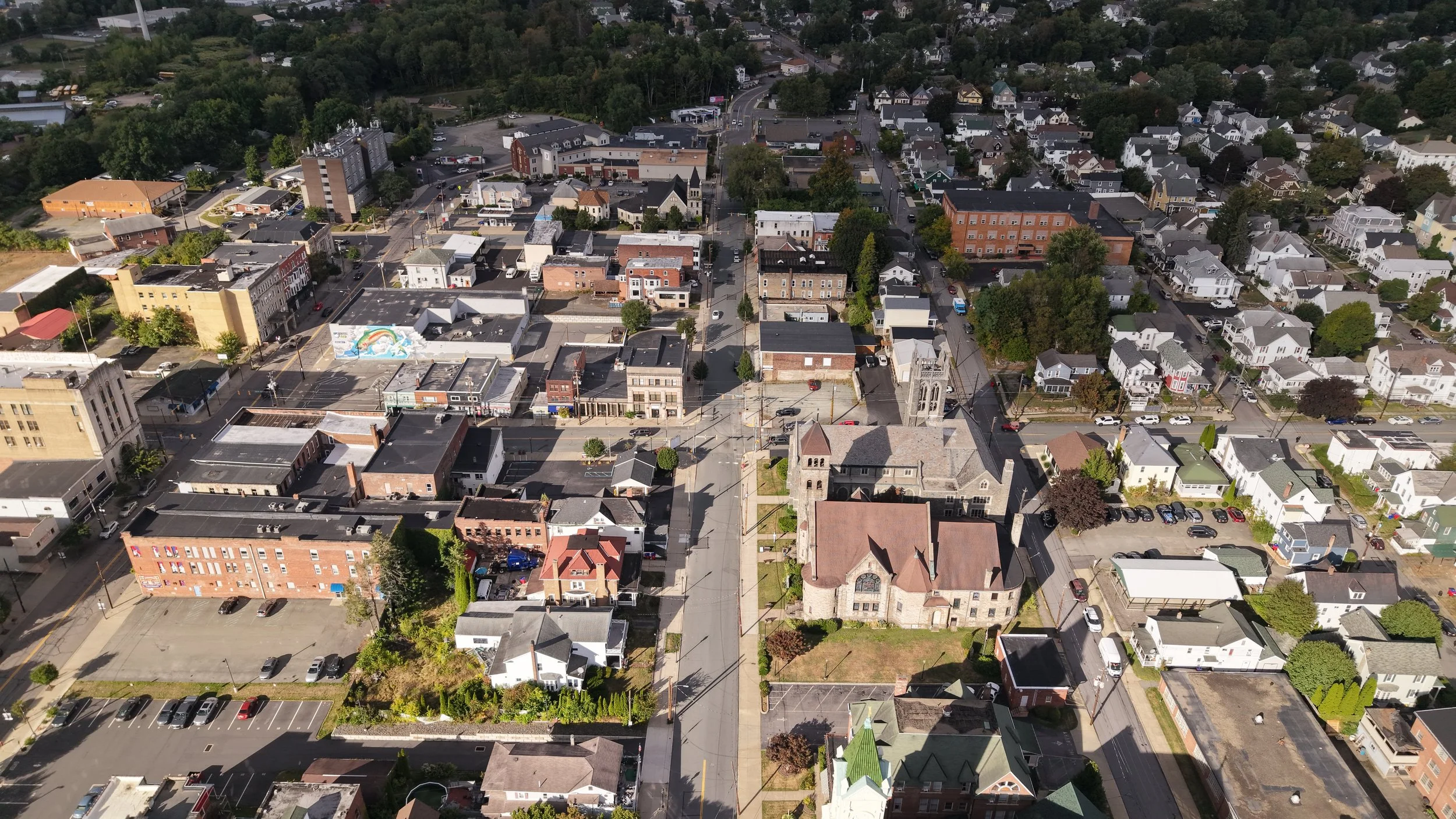 Aerial view of a small town with various buildings, streets, trees, and parking lots, surrounded by green areas and residential houses.
