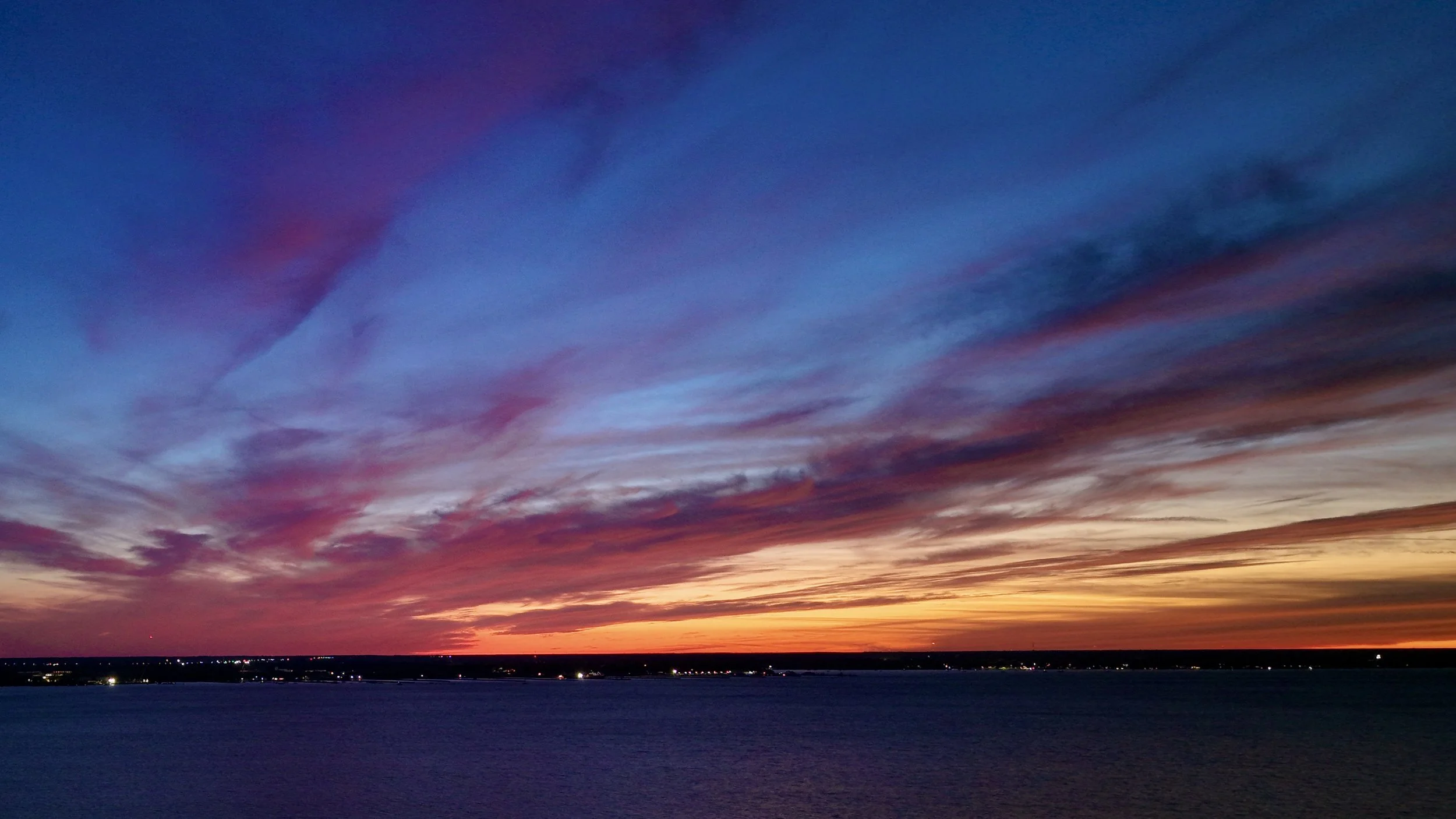 Colorful sunset over a body of water with a horizon of trees and buildings in the distance.