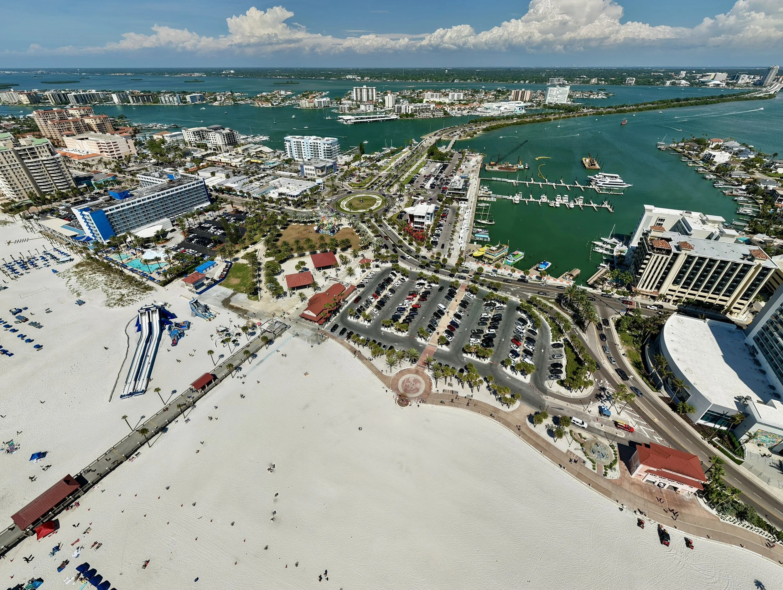 Aerial view of a city with a large sandy beach, a marina with boats, high-rise buildings, and a waterway, with clouds in the sky.