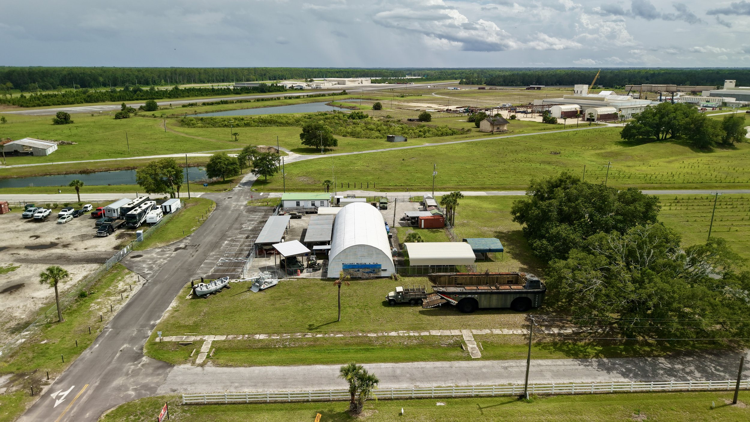 Aerial view of a rural area with open green fields, small lakes, and scattered buildings. Roads and parked vehicles are visible, along with some trees and a cloudy sky.