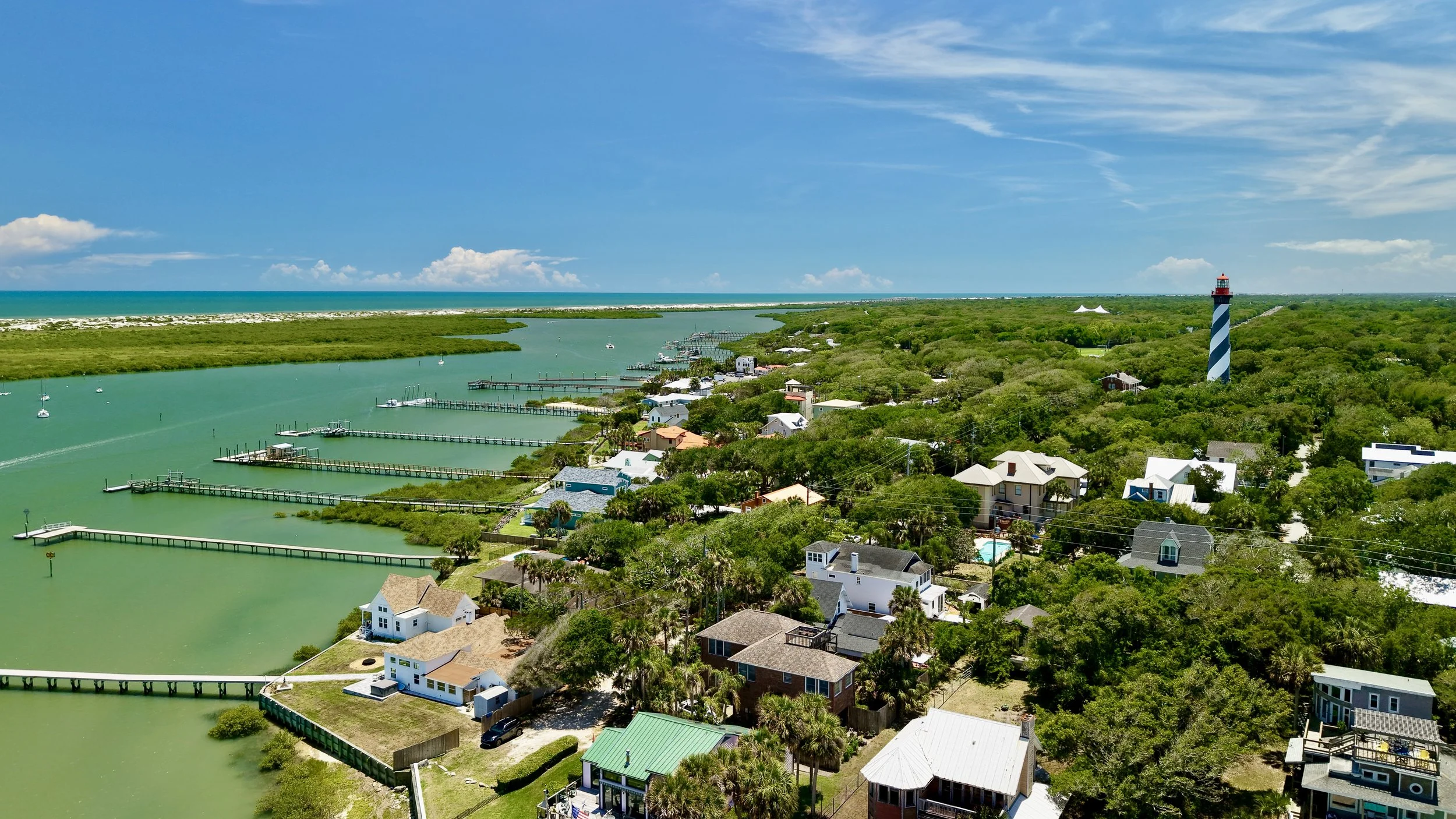 Aerial view of a coastal neighborhood with houses surrounded by greenery, a lighthouse with blue and white stripes, a waterway with boats, and the ocean in the background under a blue sky with clouds.
