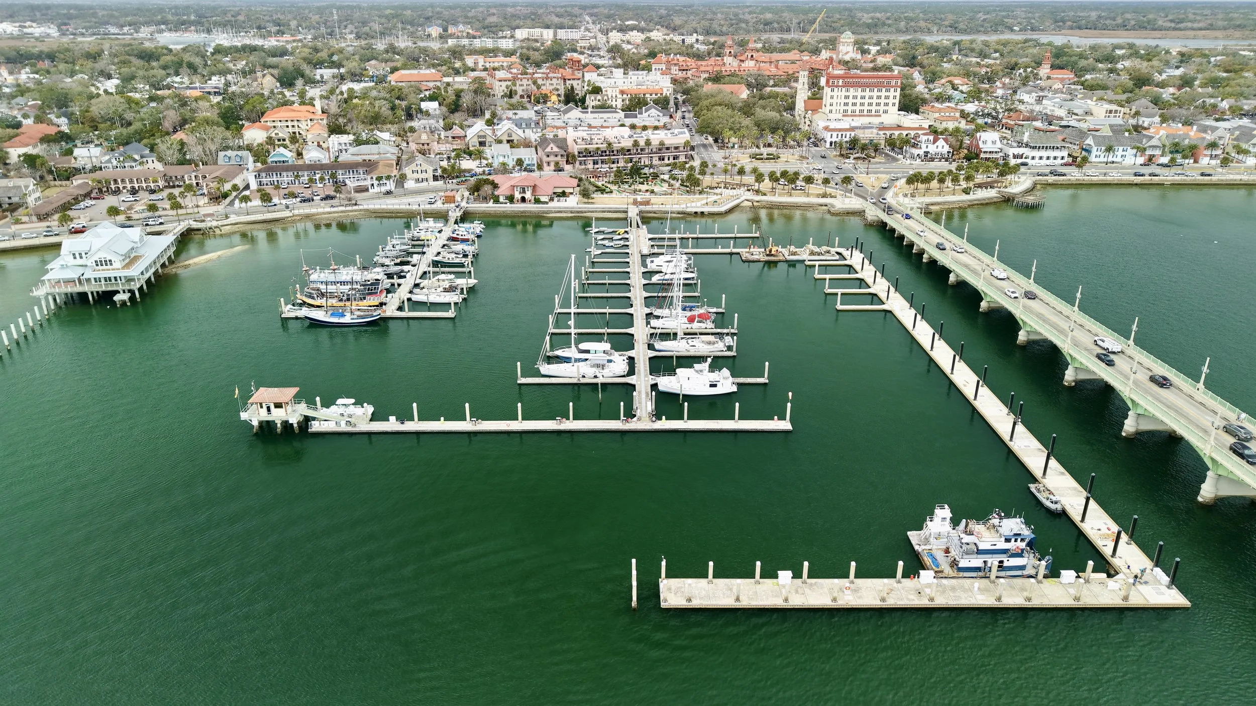 Aerial view of a marina with boats docked at piers, surrounded by a coastal town with colorful buildings, roads, and greenery.
