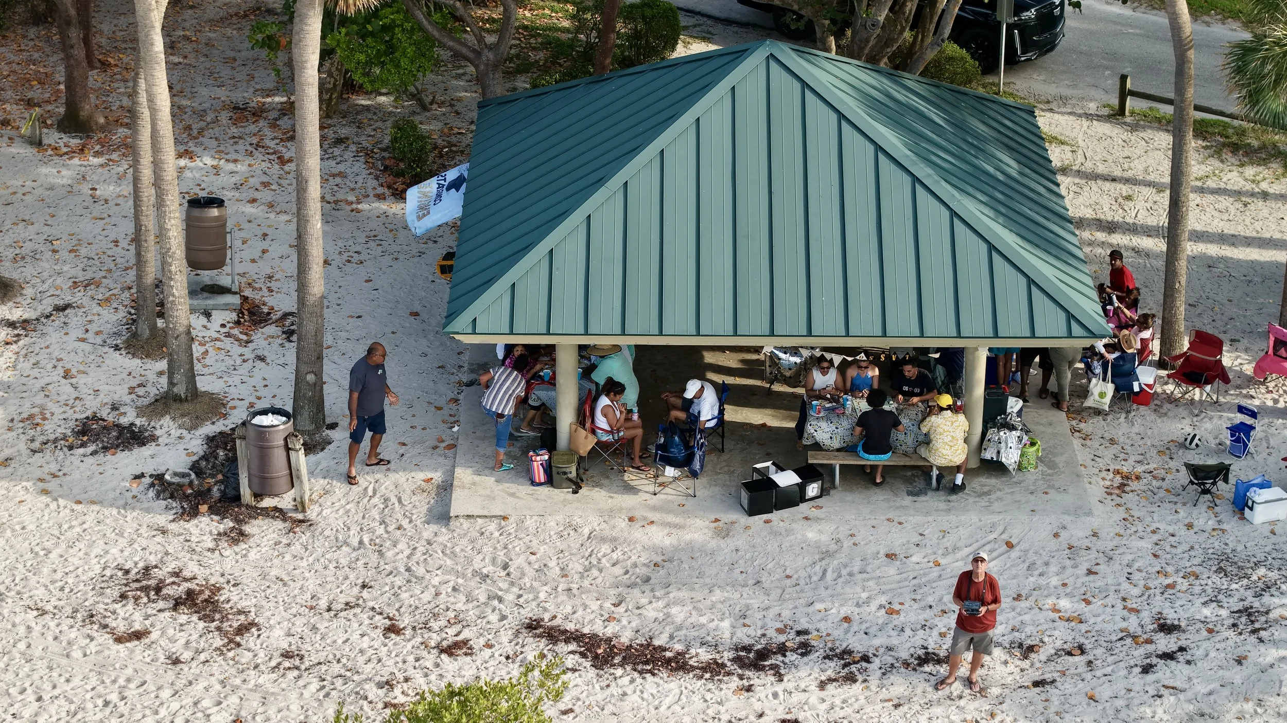 A group of people gathered under a pavilion with a green metal roof on a sandy beach. Some are sitting at table with food and drinks, while others are standing or walking around. There are umbrellas, chairs, and various beach accessories around the a