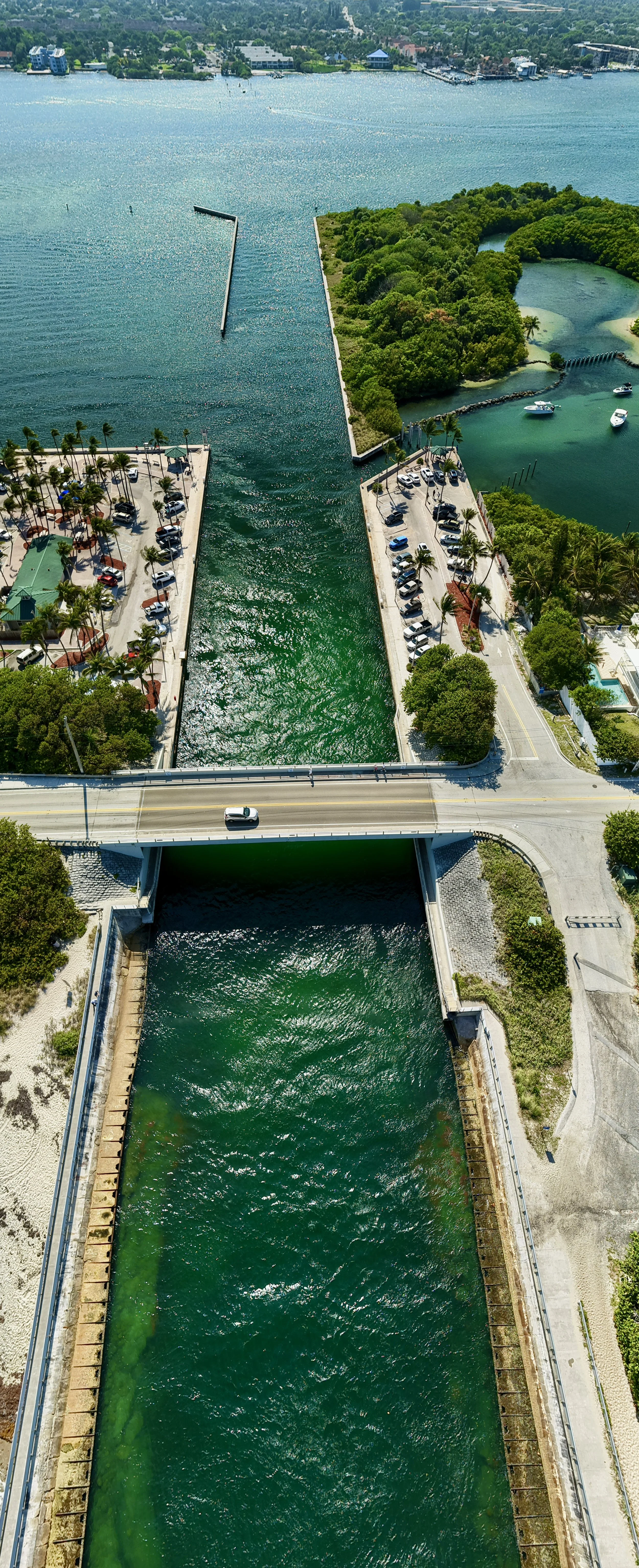 An aerial view of a waterway passing under a bridge, with two dams on either side, one with greenish water and the other with blue water. There are parking lots filled with cars, palm trees, and surrounding green areas along the water.