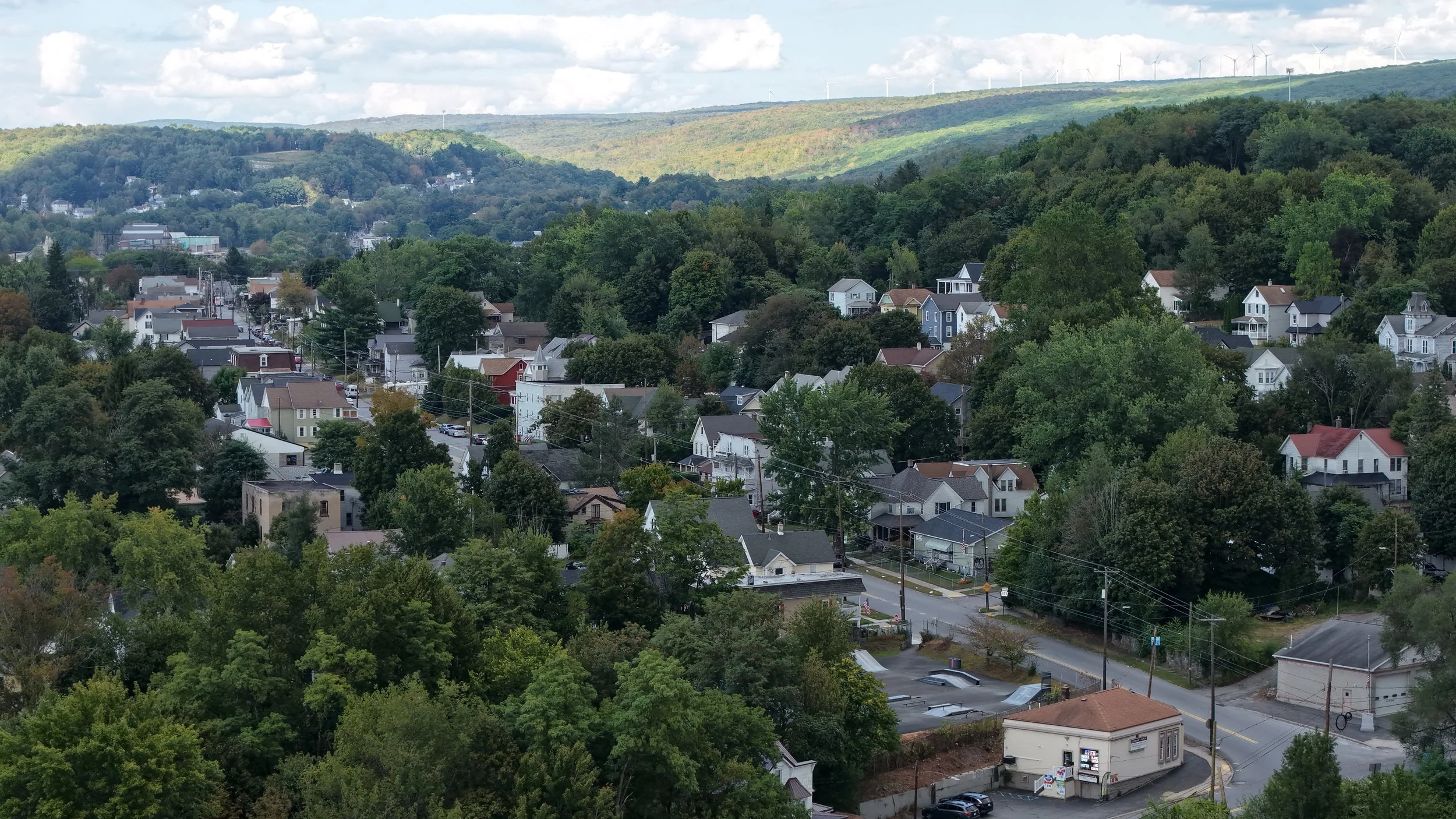 A hillside town with many houses surrounded by dense green trees and hills, some wind turbines visible on the distant hilltop.