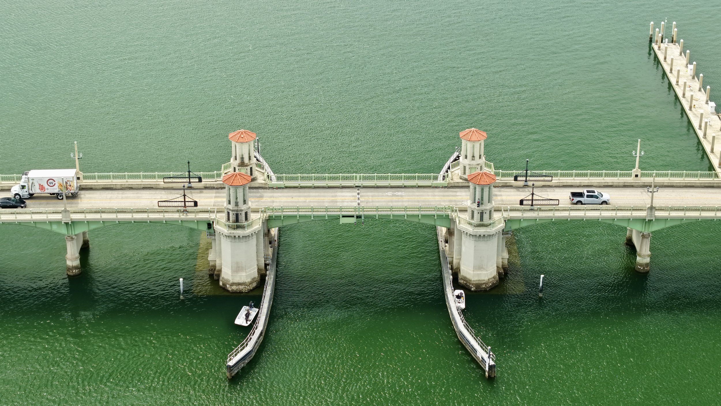 Aerial view of a bridge with sea water underneath, featuring four towers with red roofs, cars, and a boat.