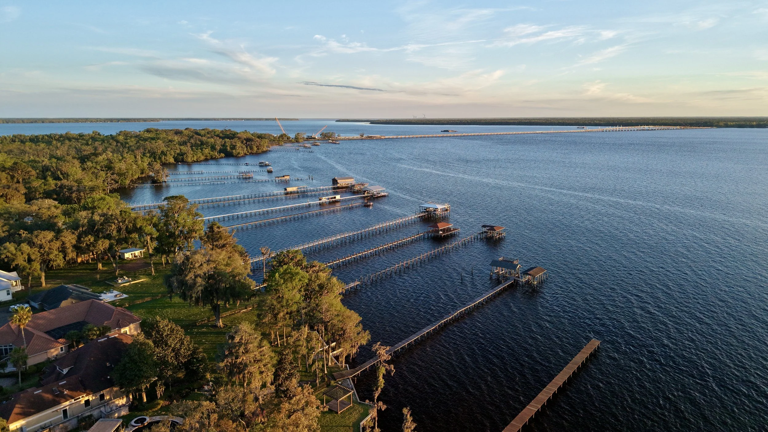 Aerial view of a lake with houses and docks along the shoreline, trees surrounding the area, and a long bridge or causeway extending across the water in the distance under a partly cloudy sky.