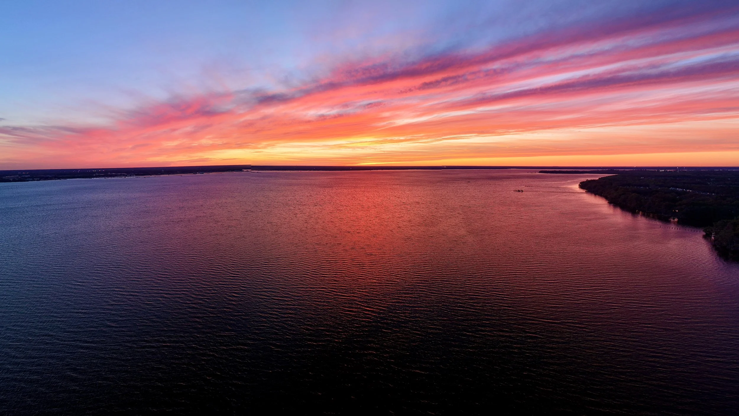 A vibrant sunset over a large body of water with colorful pink, orange, and purple clouds in the sky and a forested shoreline on the right side.