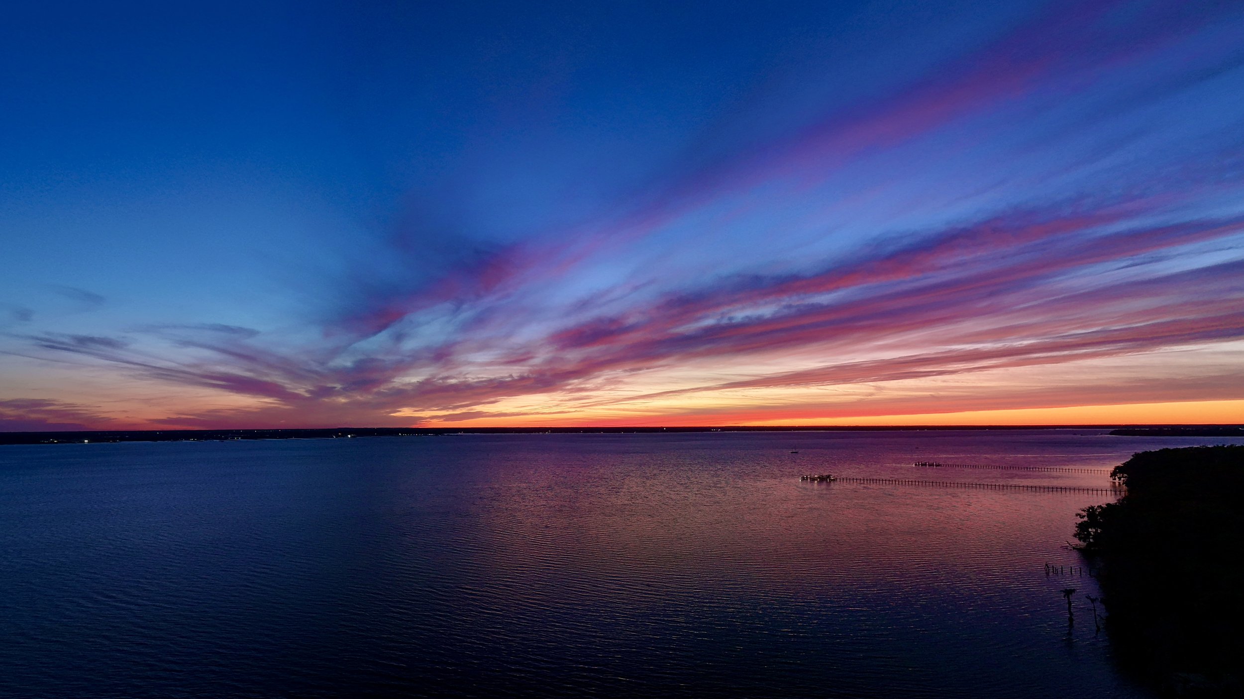 A colorful sunset over a large lake with a distant shoreline and a pier extending into the water, with silhouette trees in the foreground.