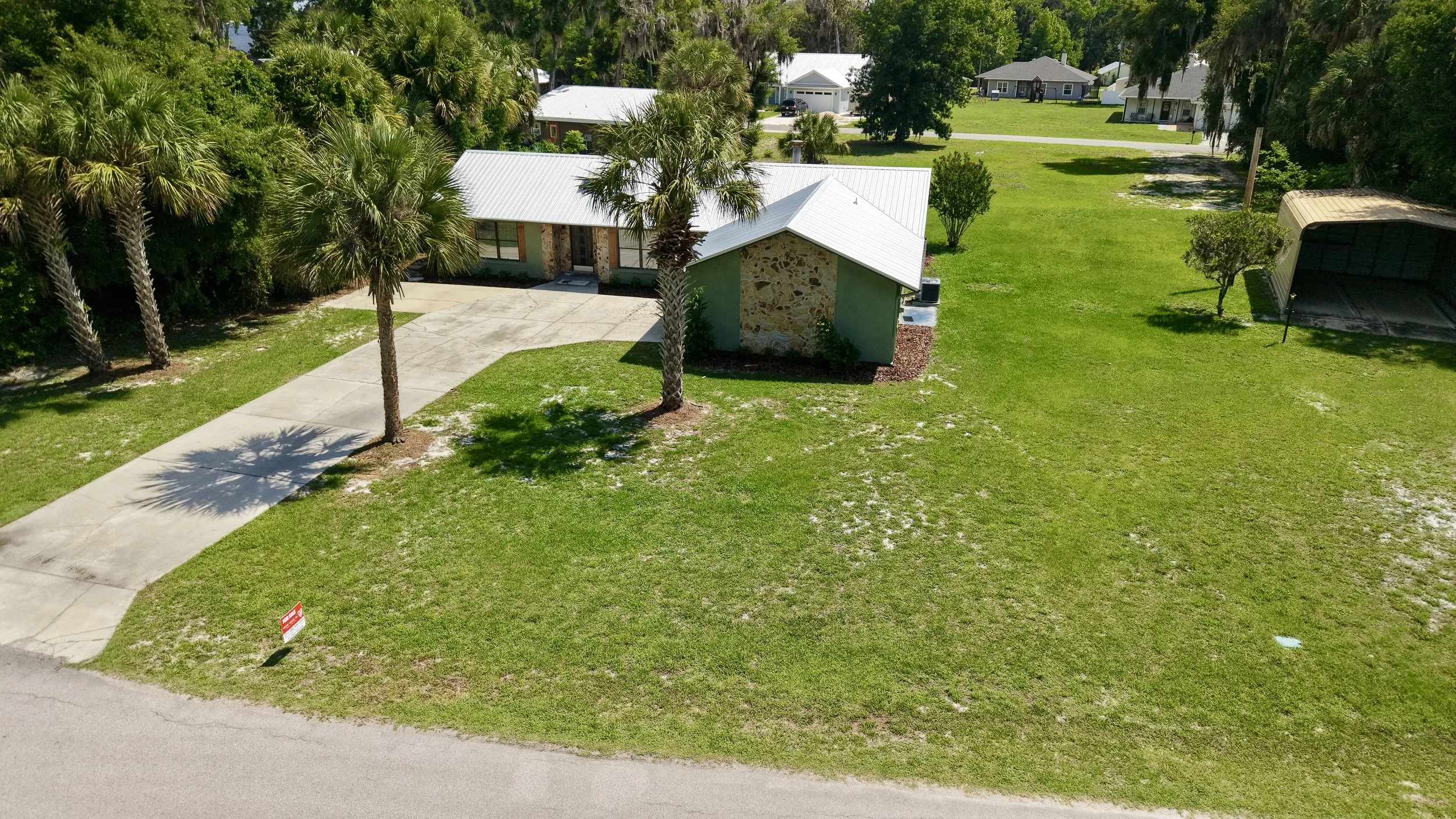 A small house with a white metal roof, stone and green exterior walls, surrounded by palm trees and grass. A concrete driveway leads to the house with an 'Inquire' sign near the street.