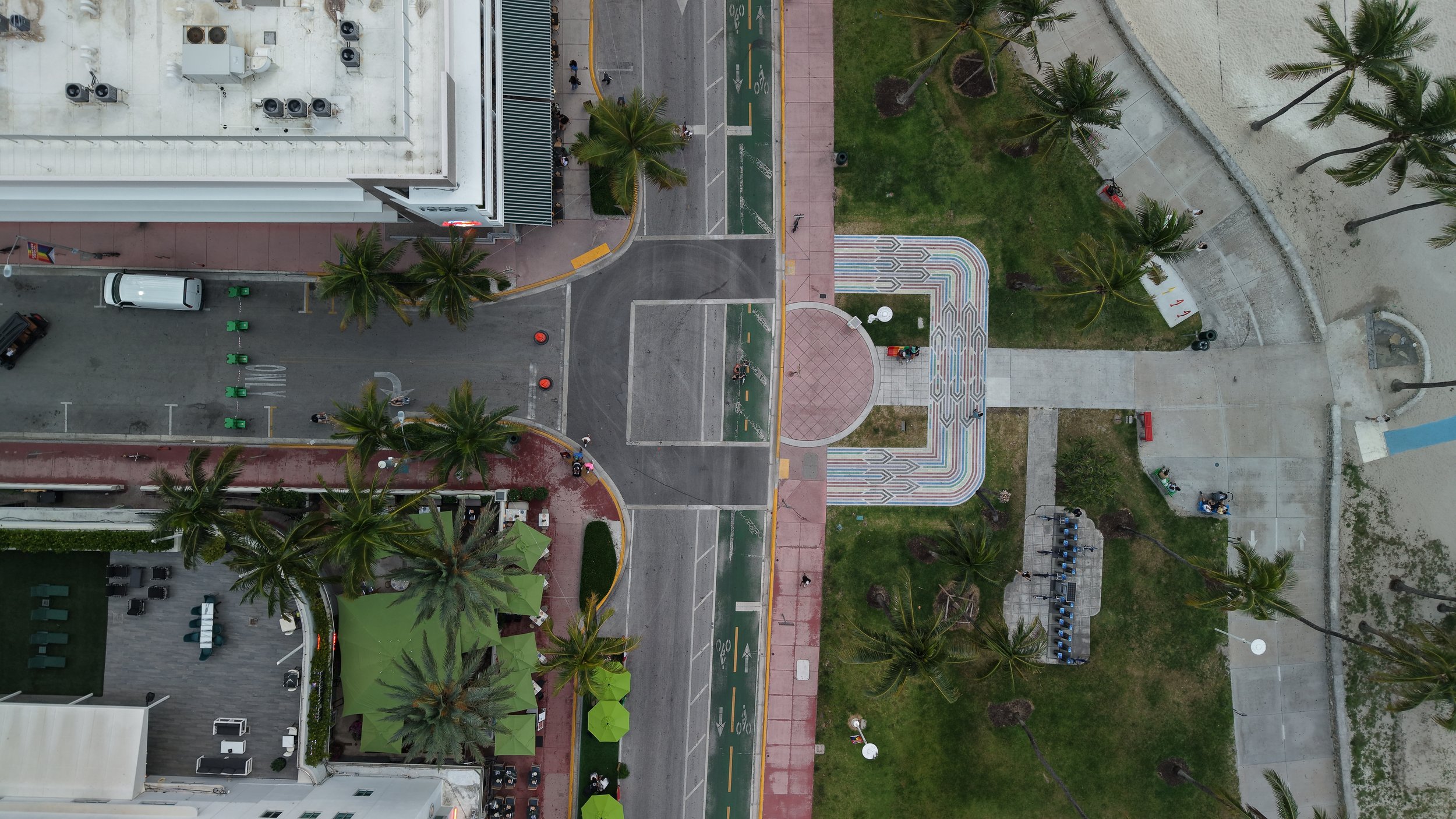 Aerial view of a city block showing a white building with air conditioning units, a parking lot with a white vehicle, a sidewalk, palm trees, a colorful graphic crosswalk, a grassy area with more palm trees, and a sandy beach with a concrete walkway.