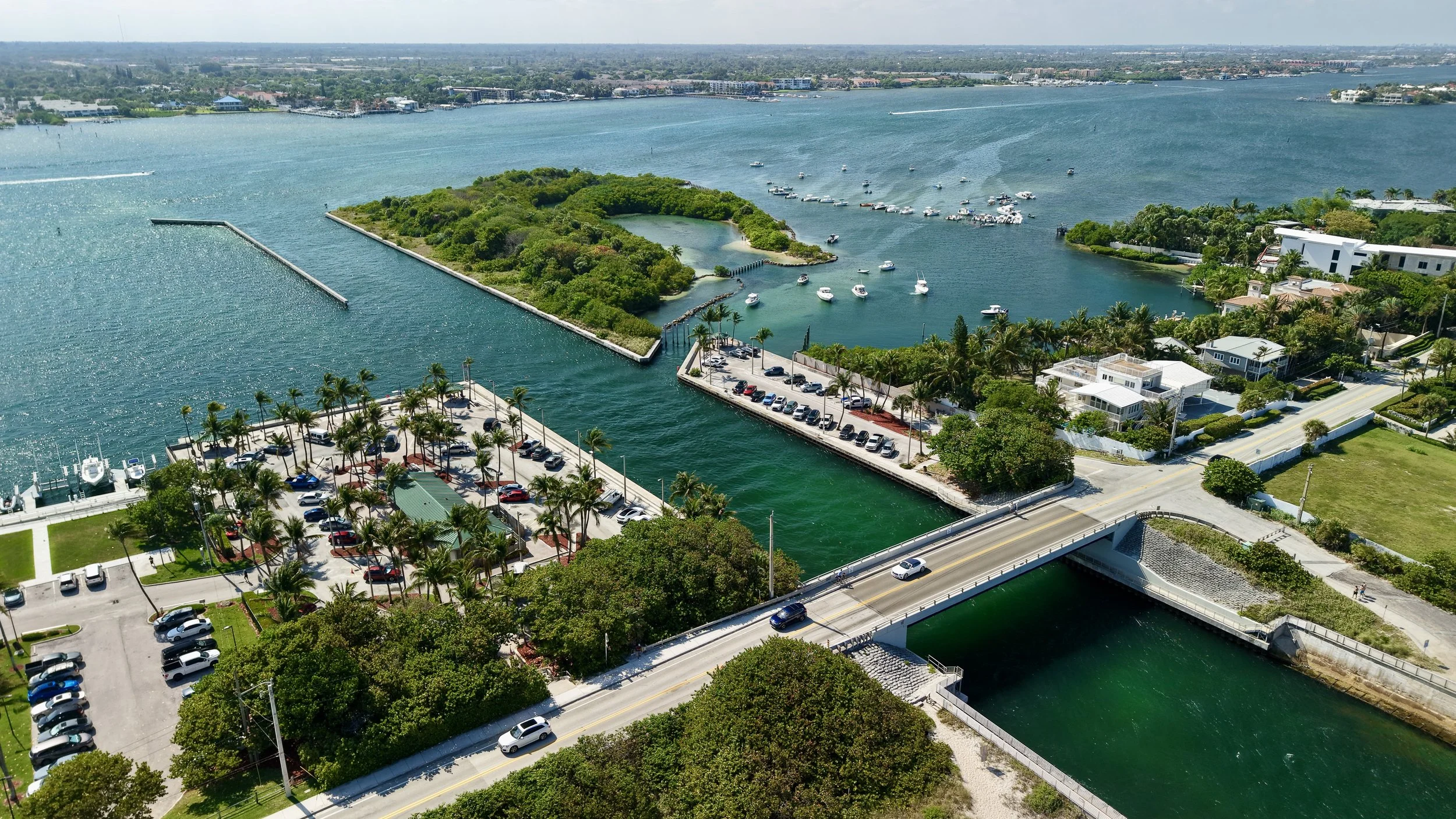 Aerial view of a coastal city with a marina, green island, and waterway, featuring boats, a bridge, and parking lots with palm trees and residential buildings.