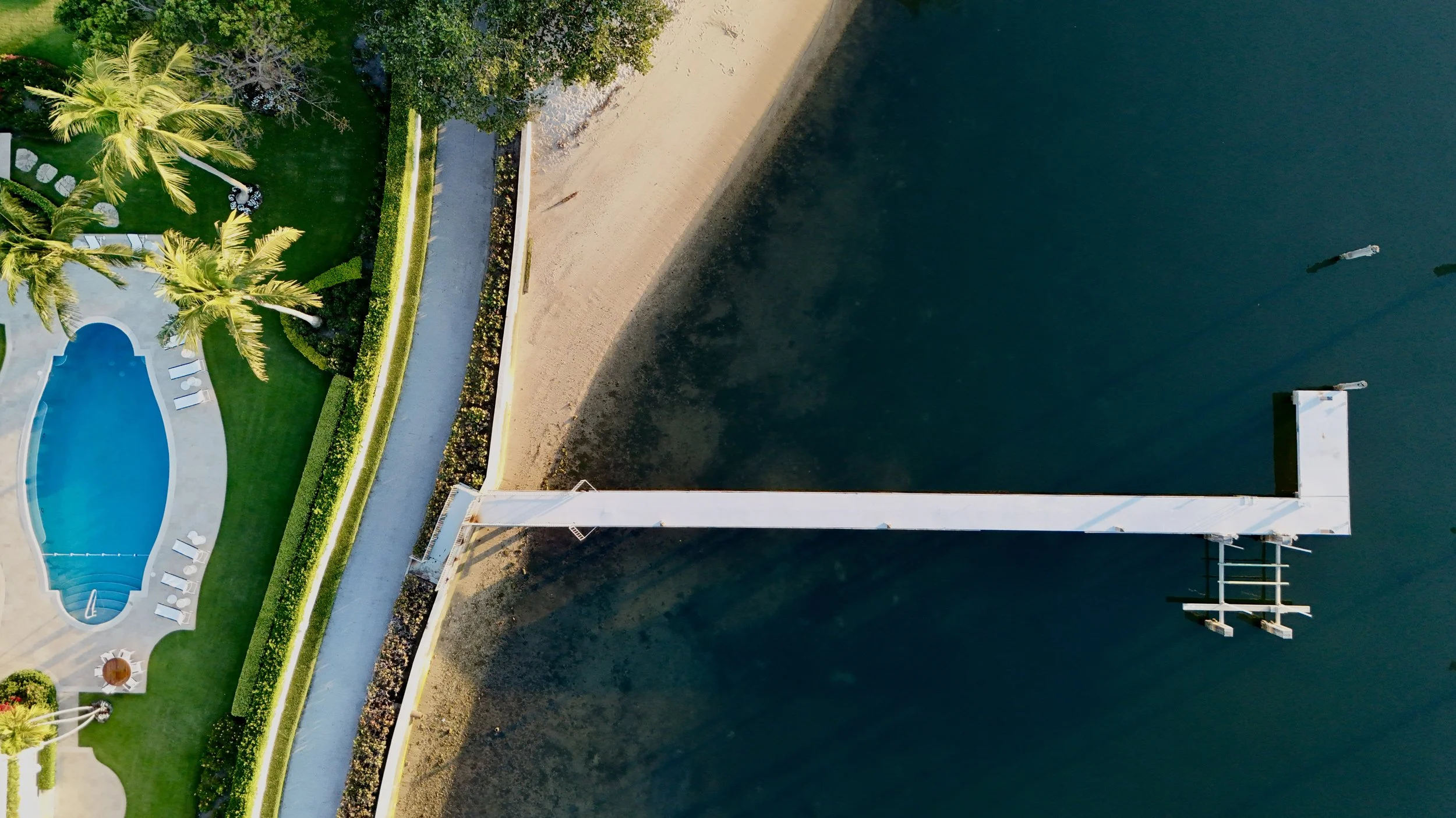 An aerial view of a waterfront property showing a sandy beach, a landscaped lawn with palm trees, a swimming pool with lounge chairs, and a dock extending into the water.