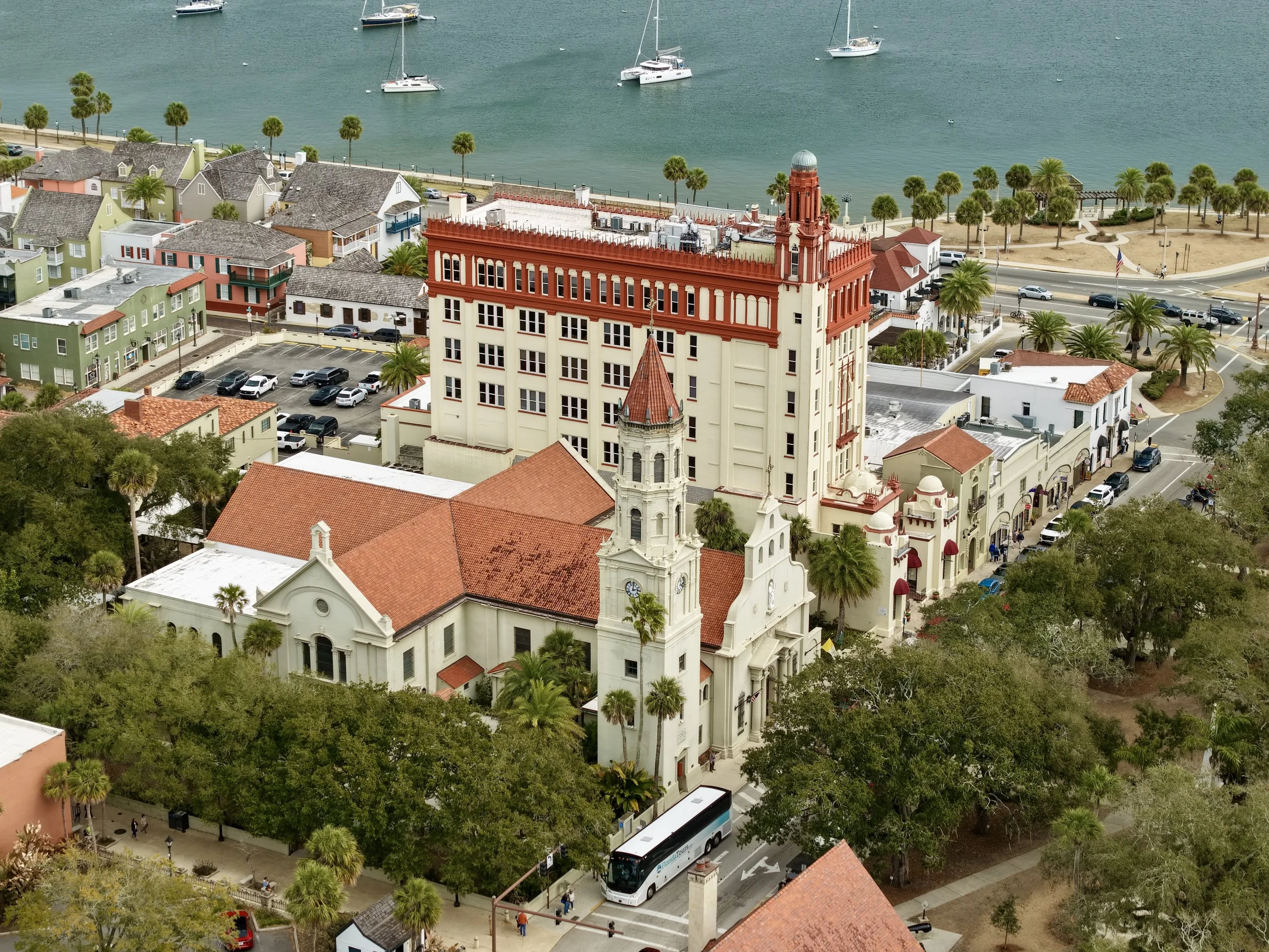 Aerial view of a coastal city with colorful buildings, palm trees, a busy street, and sailboats anchored in the water.