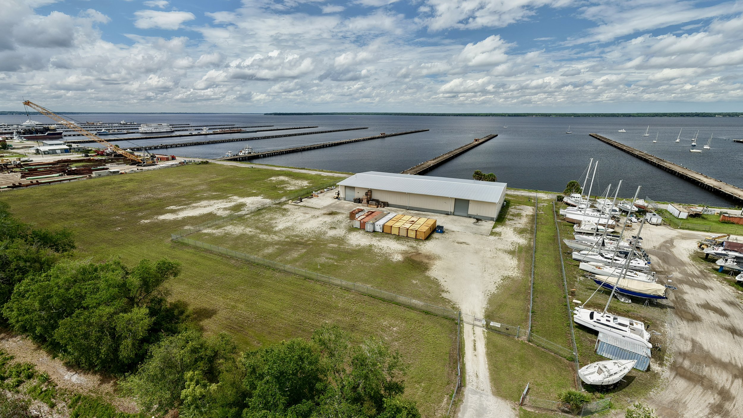 Aerial view of a marina with boats docked, a large building, storage containers, and a crane near the water on a partly cloudy day.