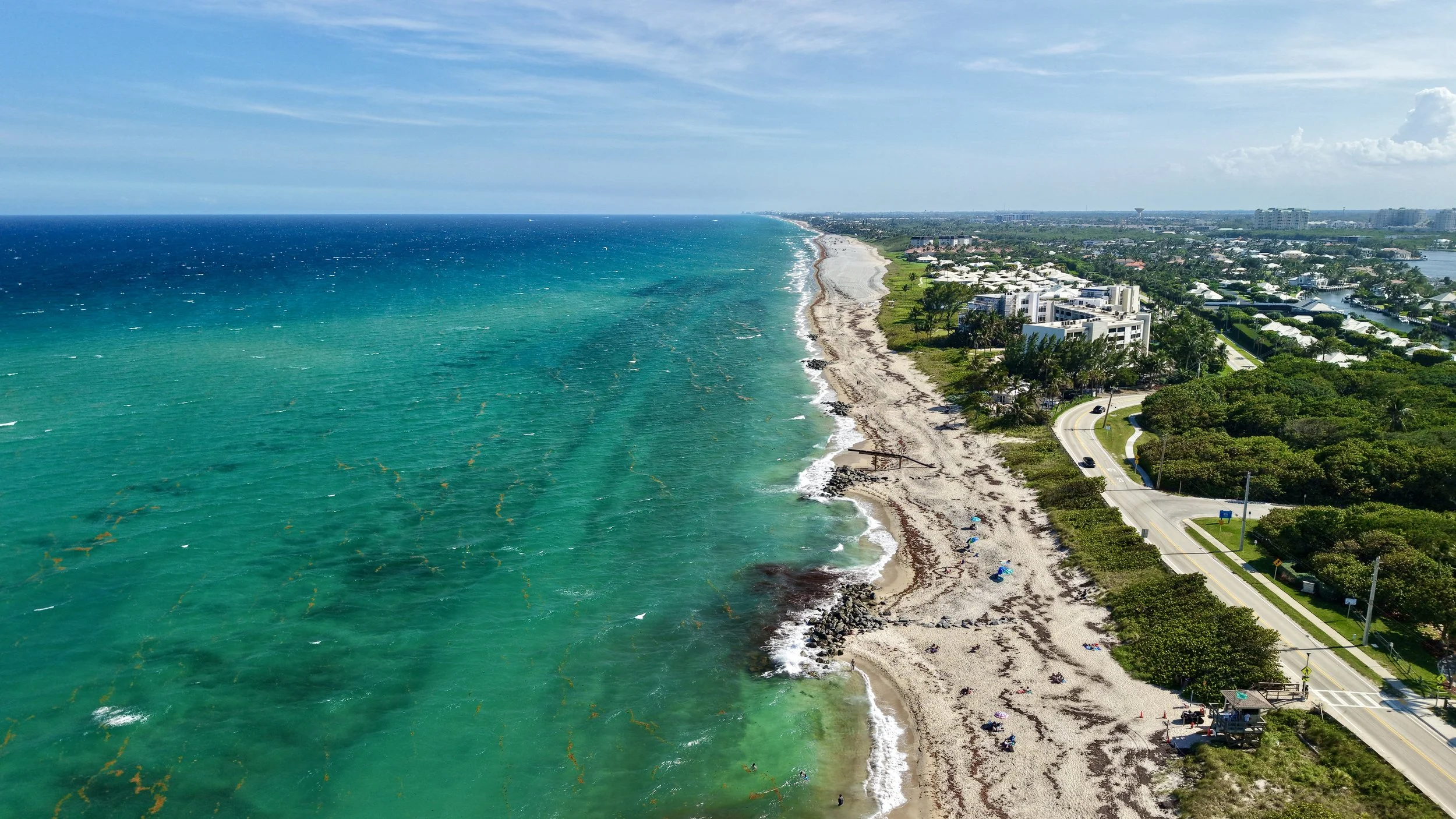 Aerial view of a coastal city with a long sandy beach, turquoise ocean water, and residential buildings along the shoreline, under a partly cloudy sky.