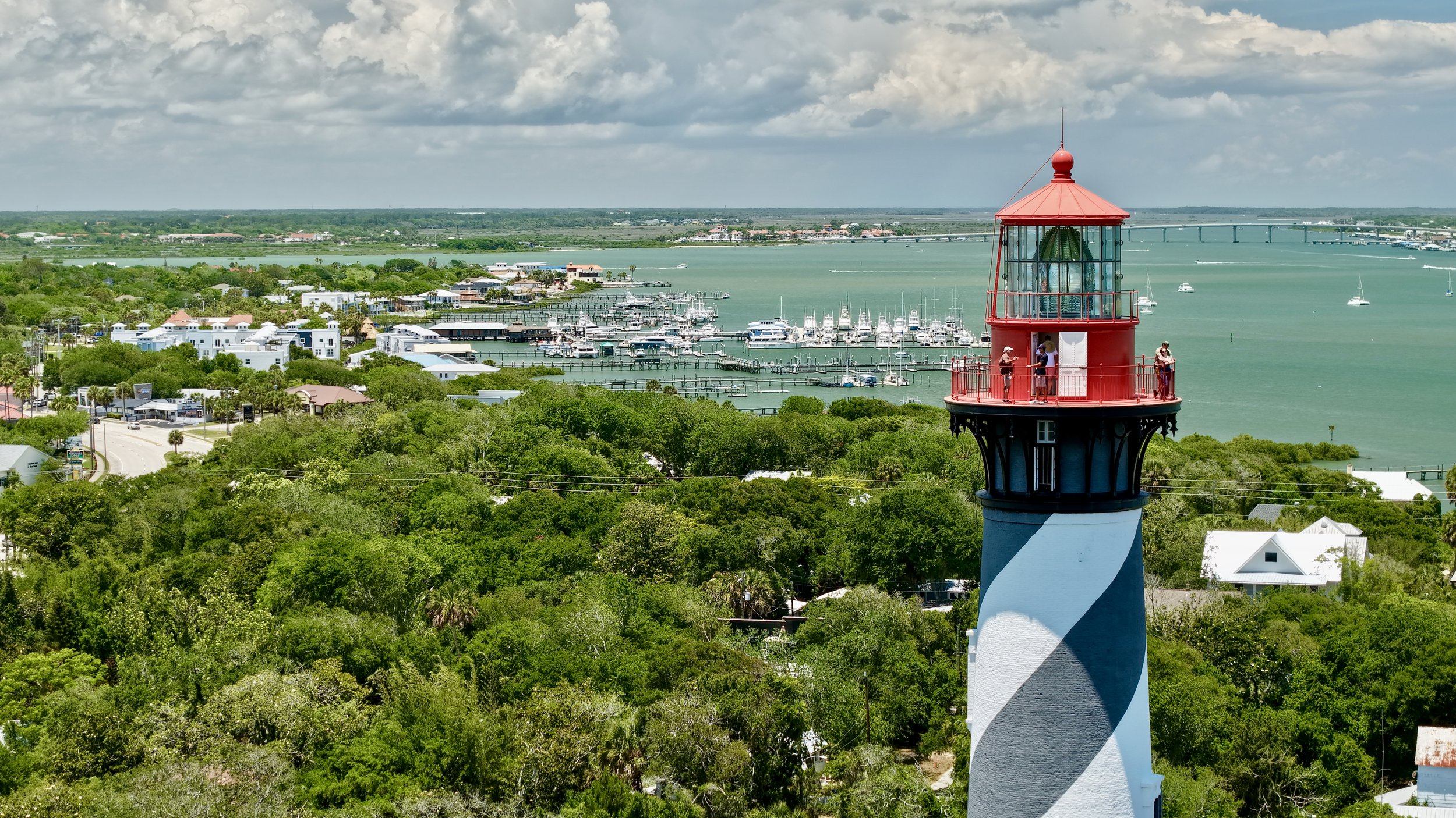 A lighthouse painted with blue and white stripes, overlooking a marina with many boats, surrounded by green trees and residential buildings, with a bridge and water in the background under cloudy skies.