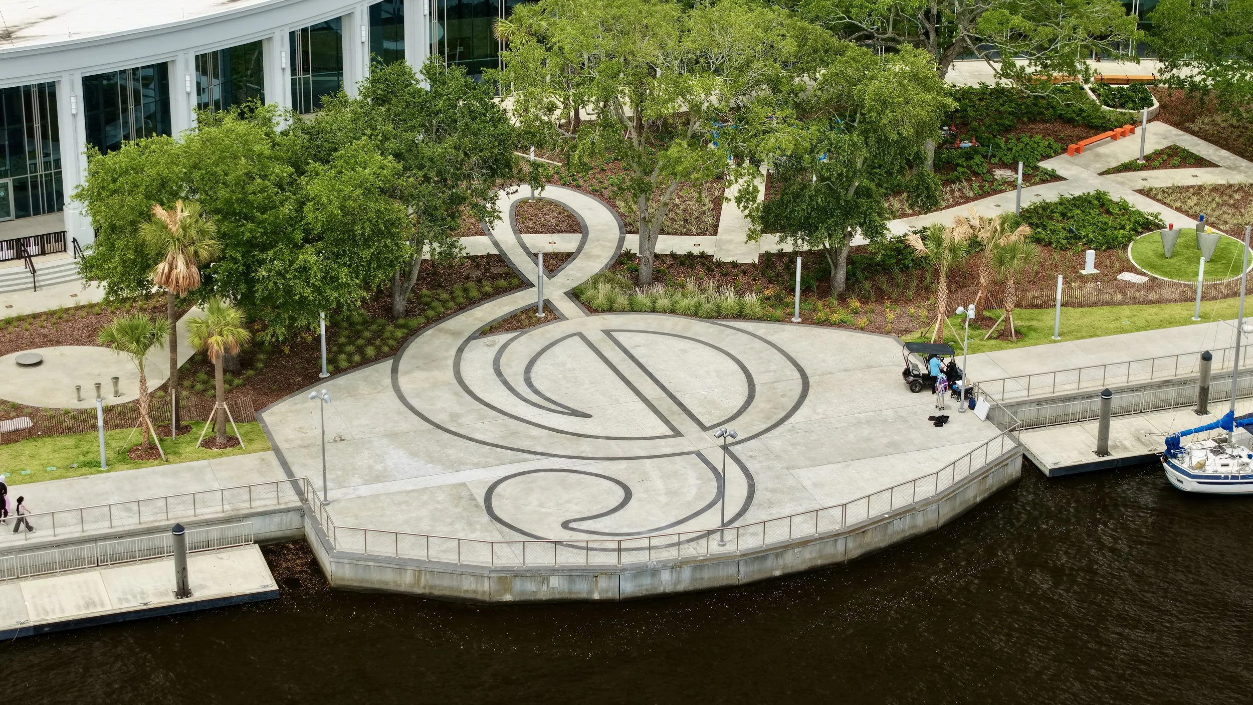 Aerial view of a landscaped outdoor area with trees, pathways, and a nautical-themed concrete design, situated beside a waterway with a docked boat.