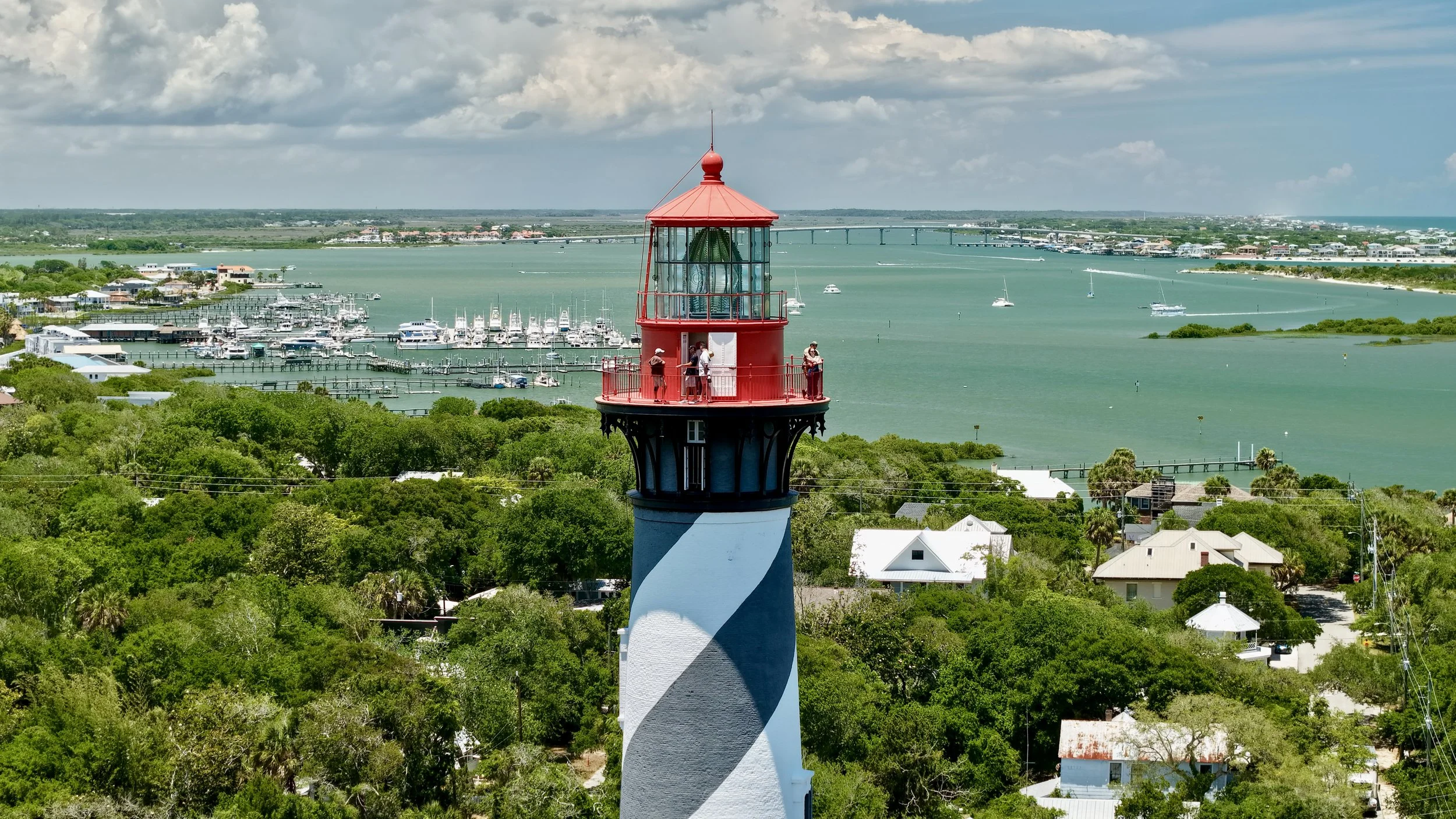 A lighthouse with a red top and white and black striped base overlooking a marina with boats, a bridge, and a distant town along a waterway under partly cloudy skies.