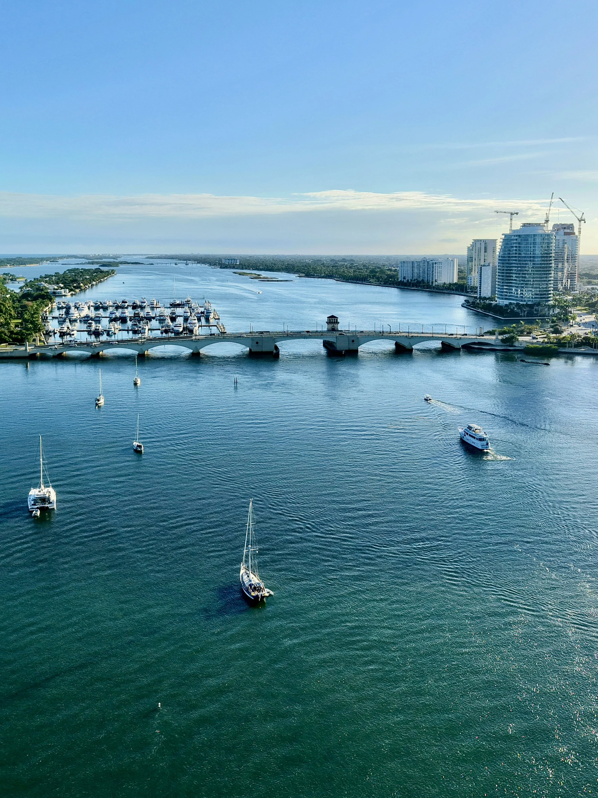 Aerial view of a river with boats and yachts, a bridge, high-rise buildings, cranes, and a clear sky.