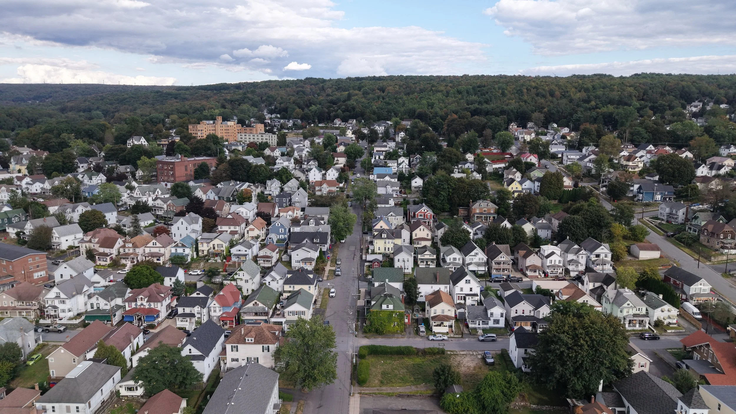 An aerial view of a residential neighborhood with a densely packed layout of houses, trees, and streets, with a forested hillside in the background.