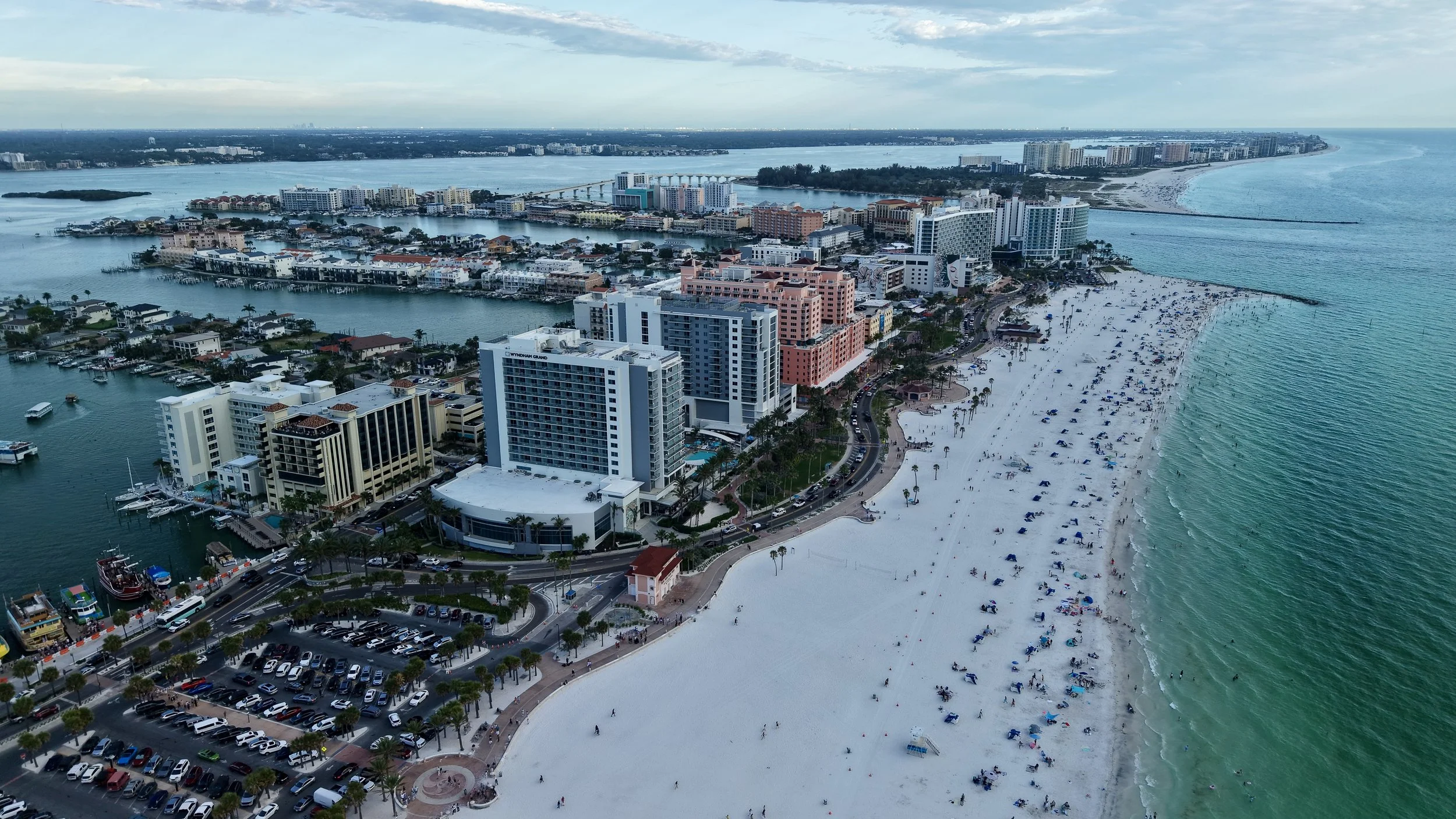 Aerial view of a coastal city with high-rise buildings, a large white sandy beach with umbrellas and people, and calm water with boats. There's a bridge connecting parts of the city across a waterway.