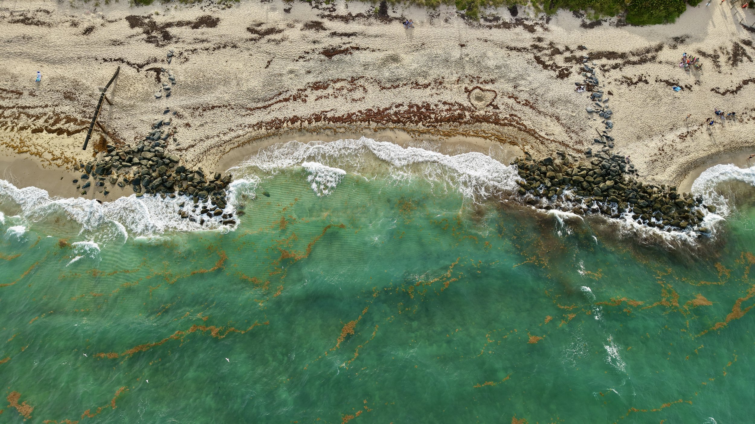 An aerial view of a sandy beach with rocks, where the words 'I love you' and a heart shape are drawn in the sand. The shoreline has waves reaching the sand, and there are some people and objects on the beach.