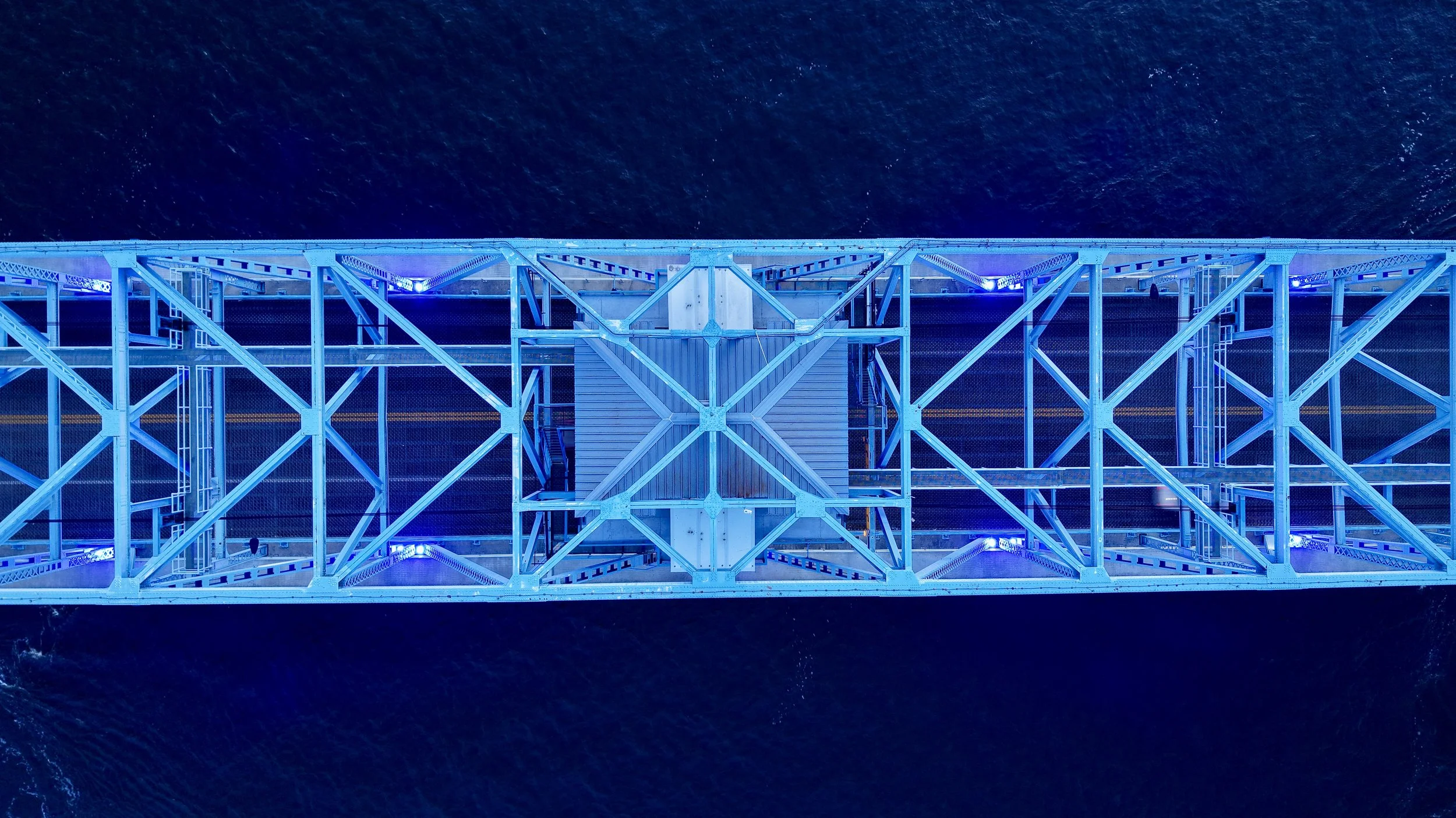 An aerial view of a bridge illuminated with blue lights, spanning over dark water.
