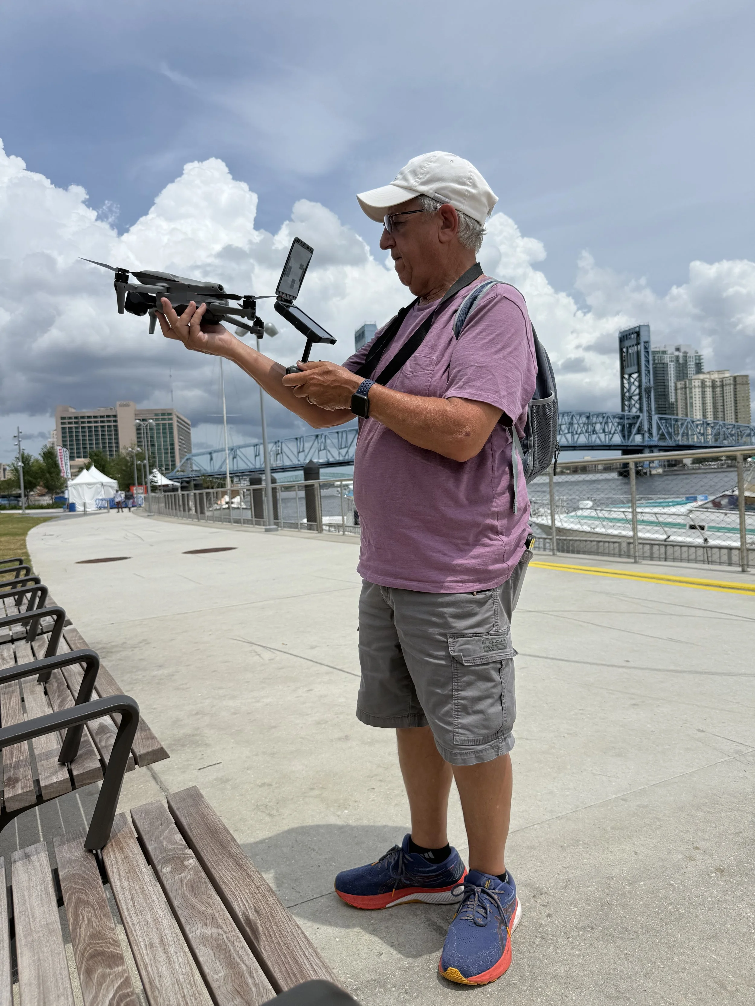 An elderly man wearing a white cap, glasses, purple t-shirt, gray shorts, and blue sneakers with red accents, standing outdoors holding a drone with a smartphone mounted on a gimbal. There are benches and a skyline with buildings and a bridge in the background.
