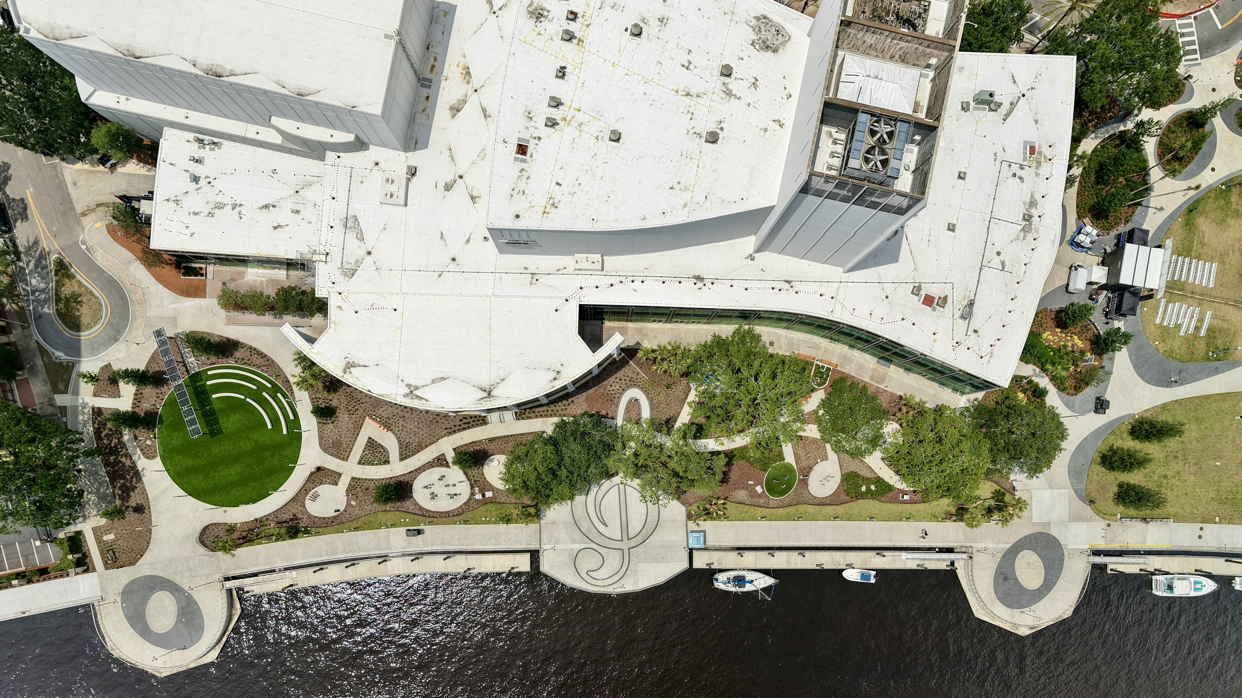 Aerial view of a modern building with a white, flat roof, surrounded by sidewalks, trees, and a waterway with boats, and landscaped areas including a green circular space with seating.