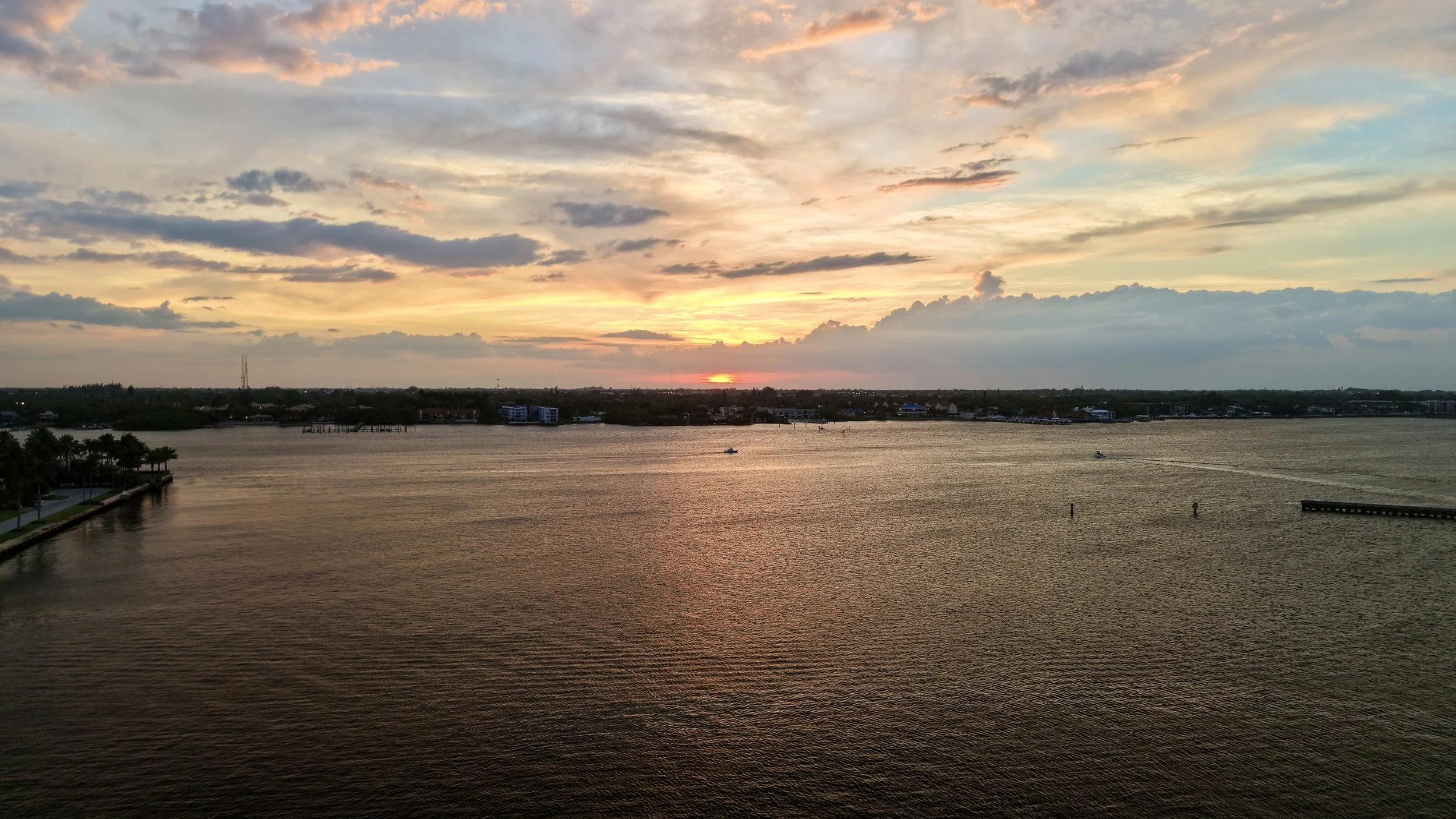 A sunset over a body of water with a colorful sky featuring clouds and hues of pink, orange, and blue. There are boats on the water, and a shoreline with trees and buildings in the distance.
