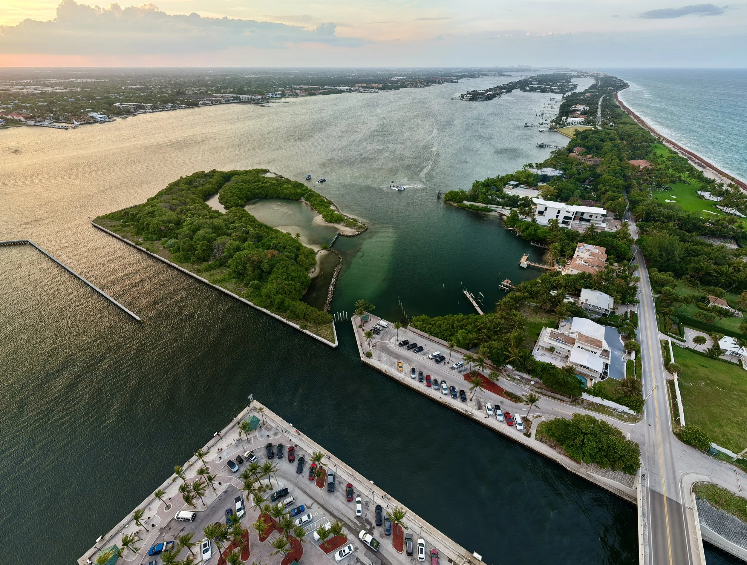An aerial view of a coastal city with a waterway, green parks, residential buildings, and a parking lot with cars. The shoreline is visible along the right side of the image, with the ocean beyond.