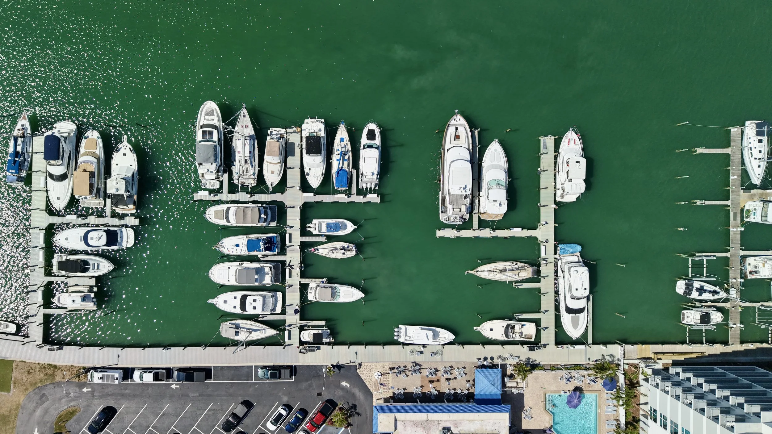 Aerial view of a marina with rows of boats docked in green water, adjacent to a parking lot and a building with a swimming pool.