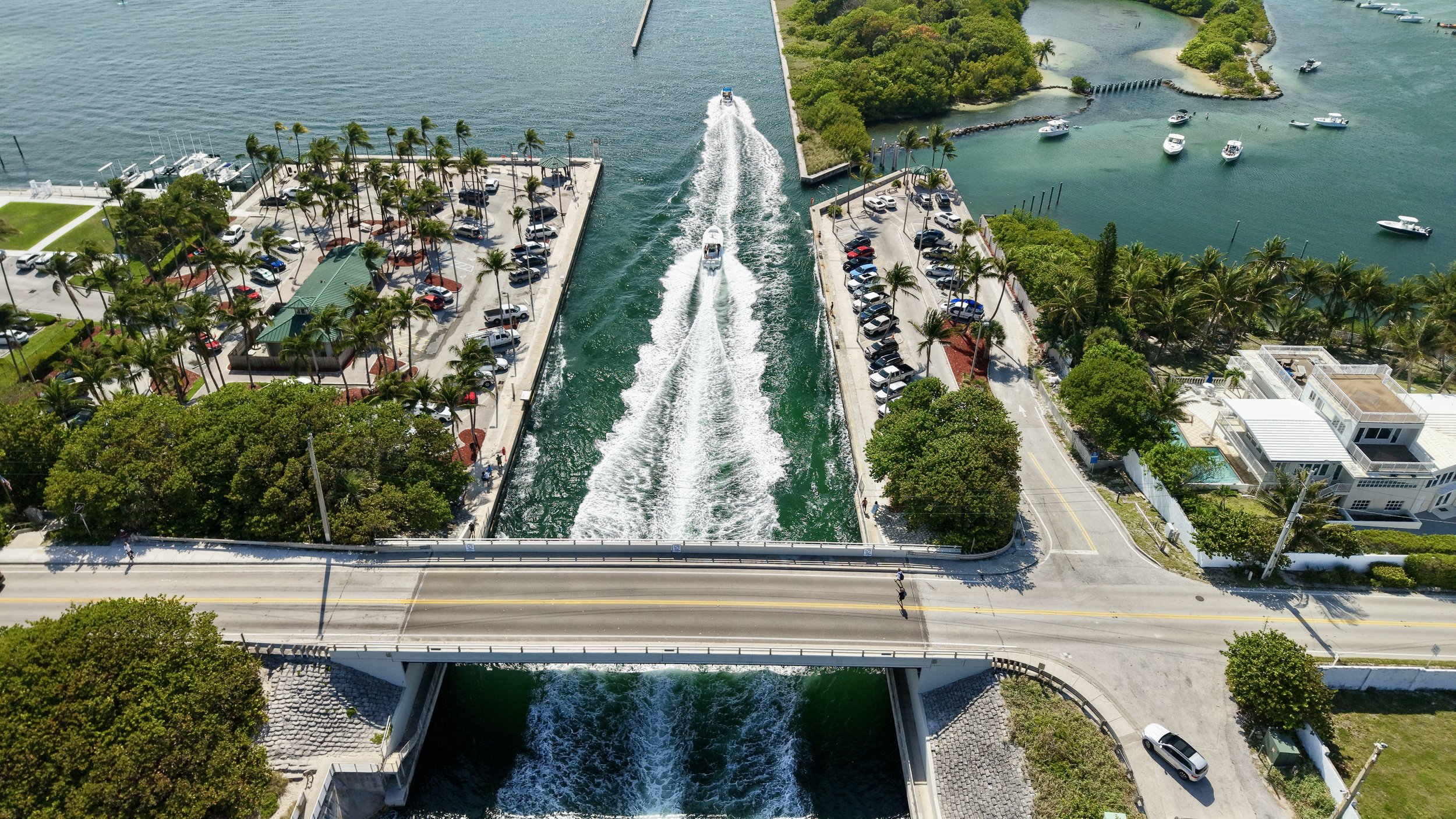 Aerial view of a waterway with boats, a bridge, and adjacent parking lots with cars. Lush greenery and buildings line the sides.