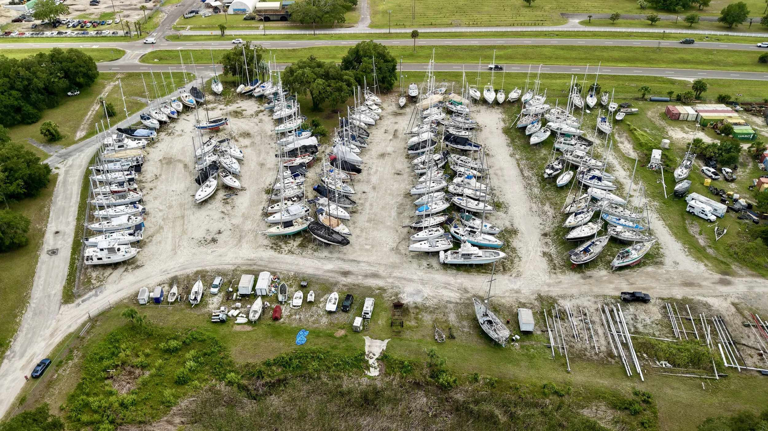 An aerial view of a boat storage yard with numerous sailboats and yachts stored on land, surrounded by green trees and adjacent to a radio station with satellite dishes.