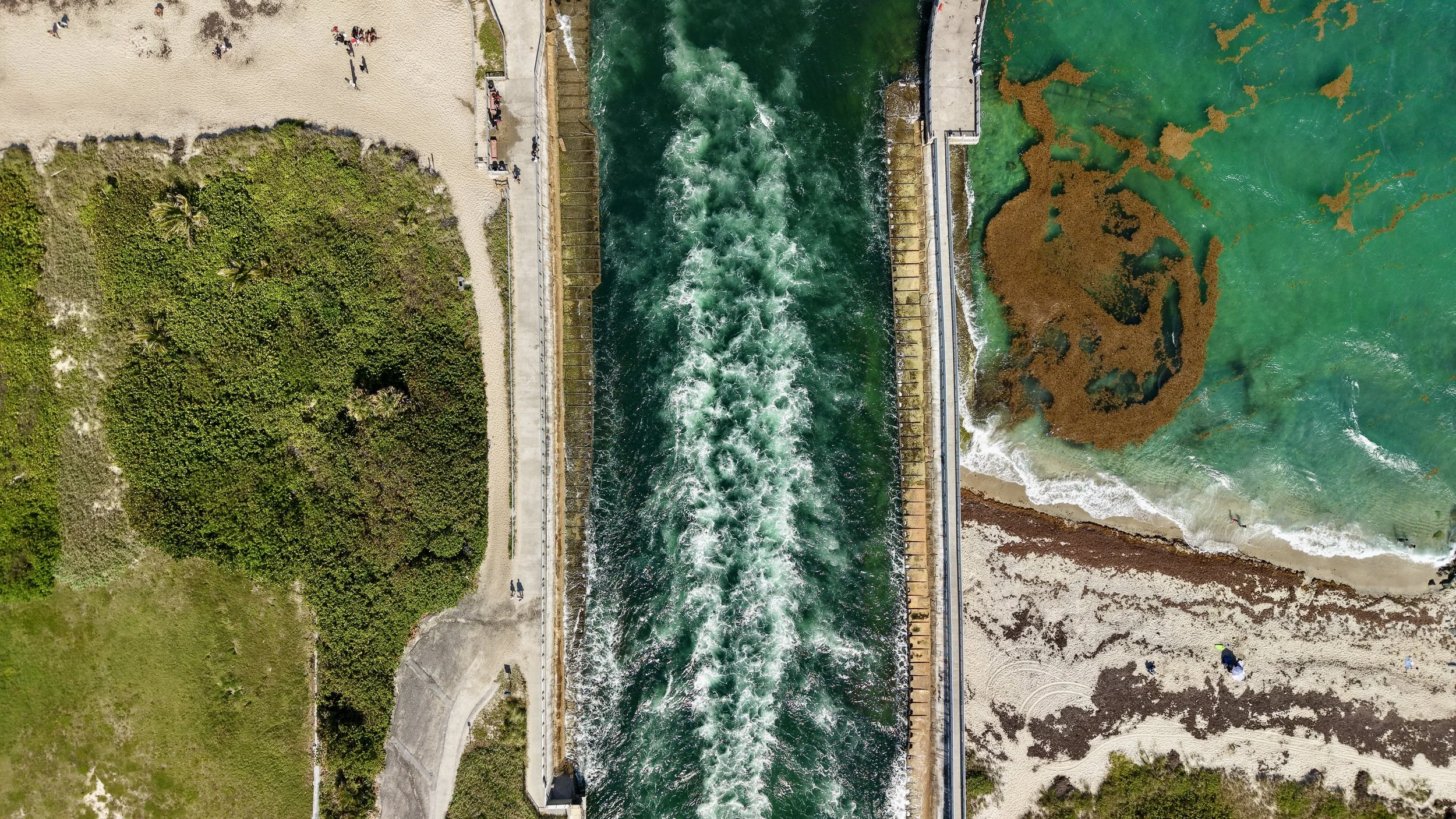 An aerial view of a dam with flowing water, sandy and grassy areas on either side, and a few people walking along the path, with the ocean visible on the right side.