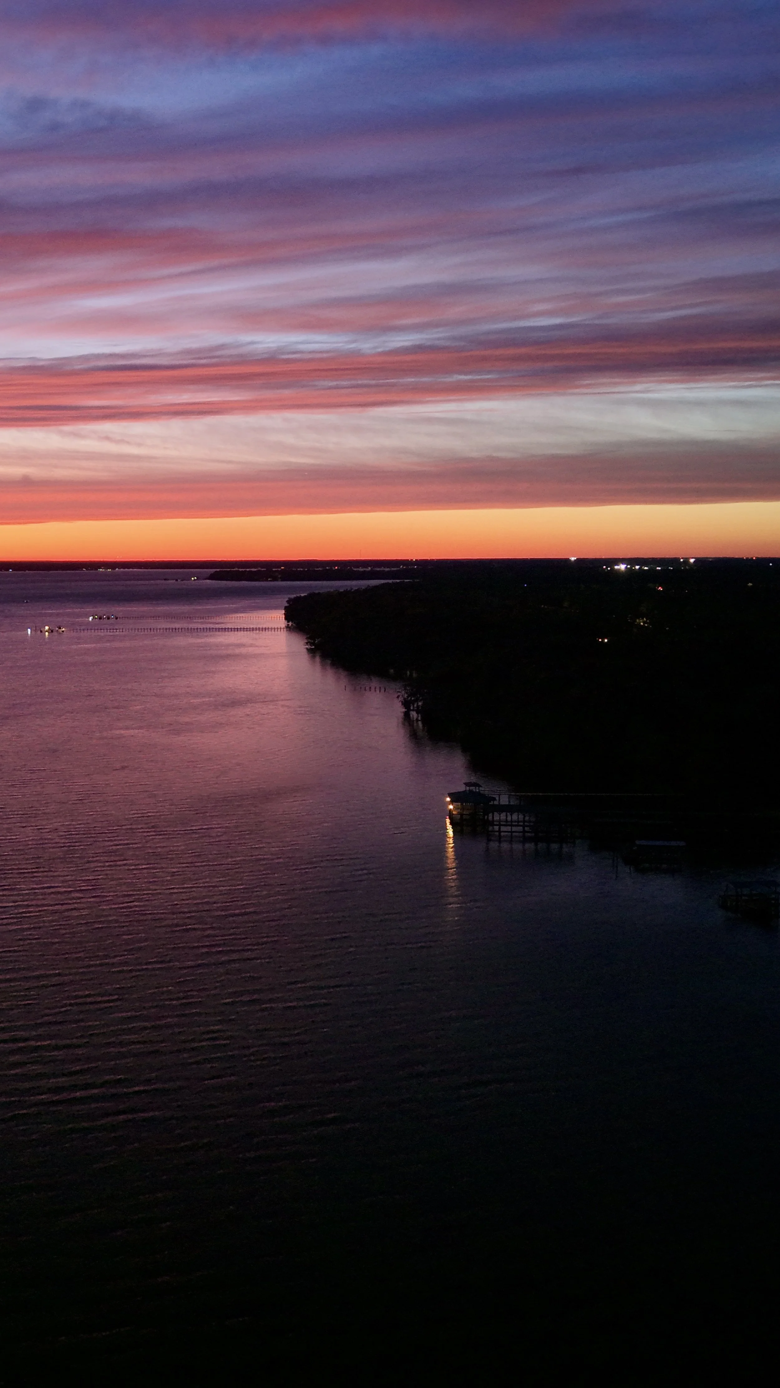 Sunset over a river with colorful sky clouds, a pier with a small shelter, and some lights on the horizon.