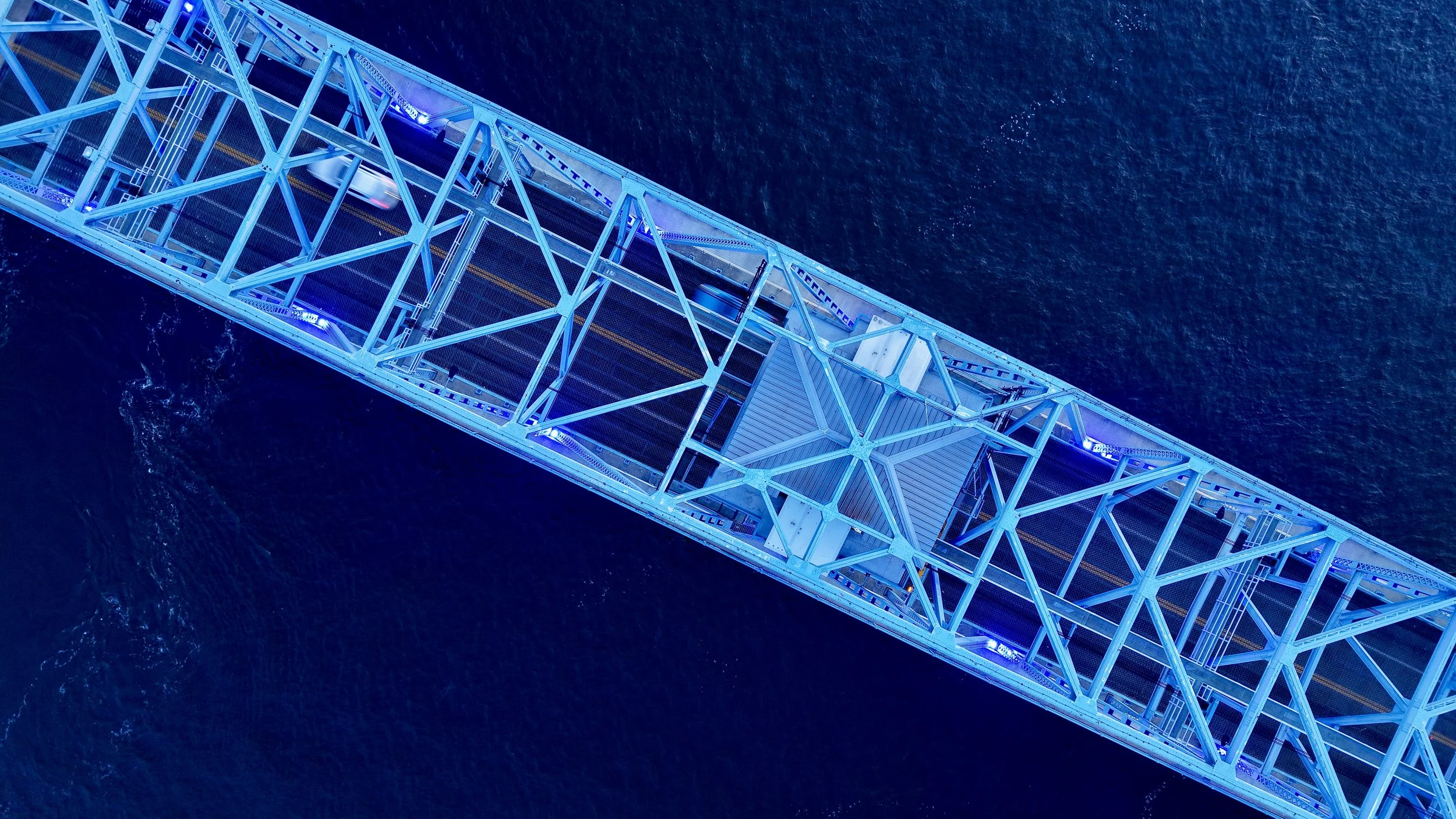 Aerial view of a bridge with steel framework illuminated in blue, spanning over a body of water.