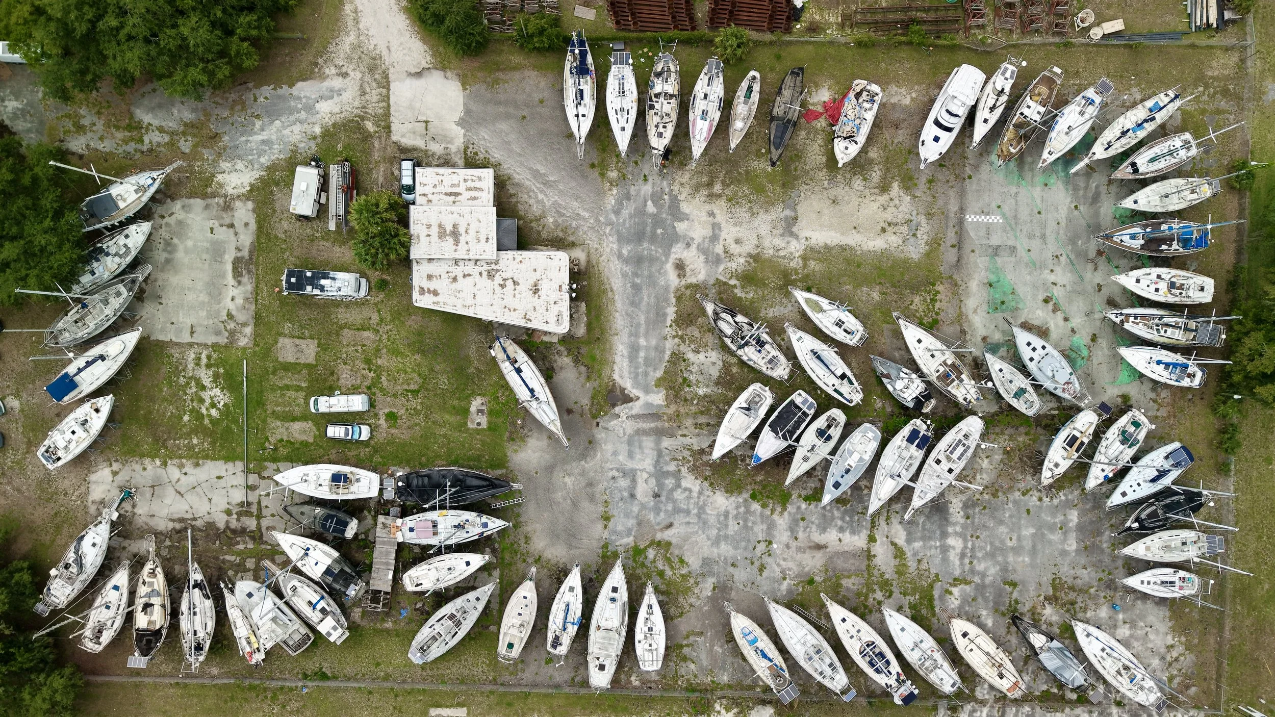 An aerial view of a boat storage area with numerous sailboats parked on land, some with sails down, and a few cars and small structures nearby, surrounded by green trees.